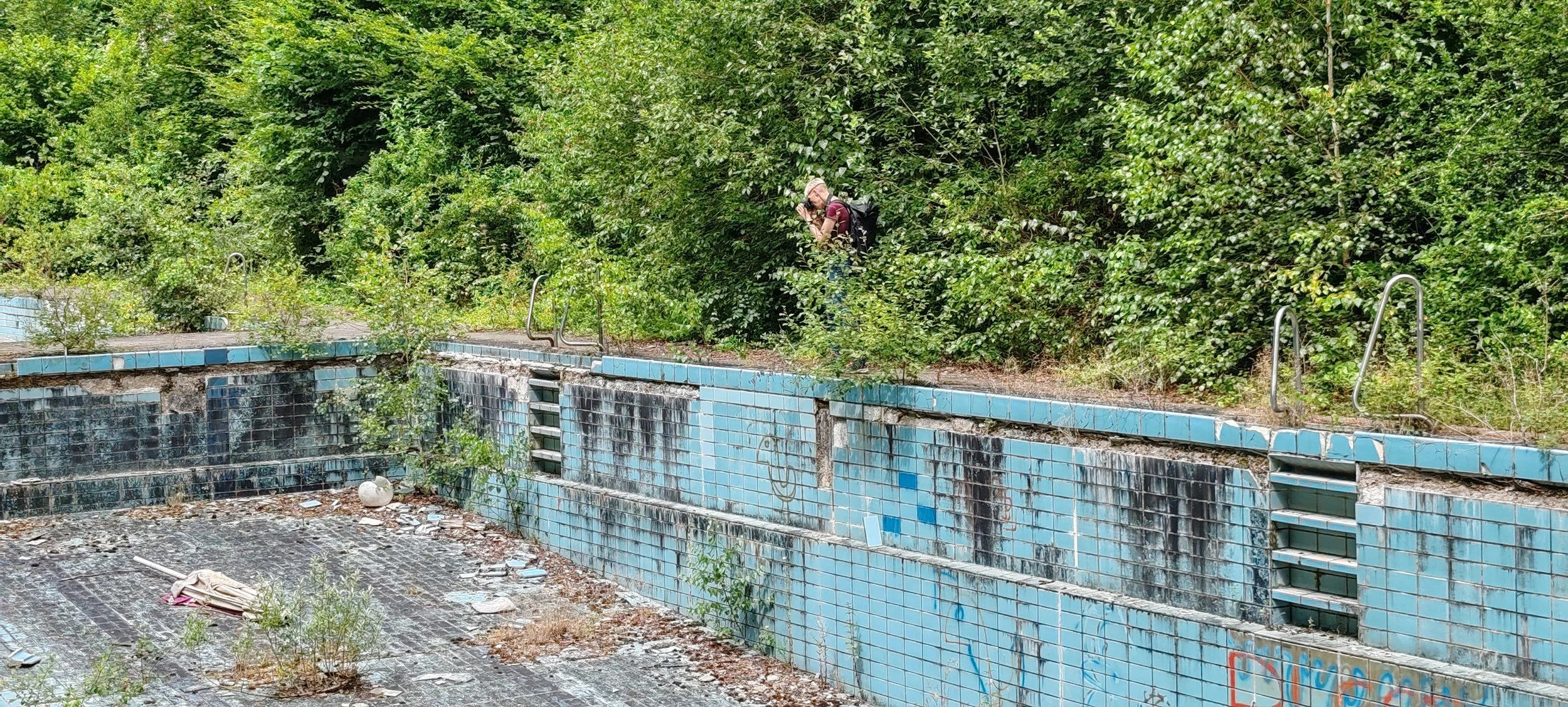 Verlassener, verwahrloster Swimming-Pool mit verwitterten blauen Fliesen und Überwucherung durch Pflanzen, mit einem Mann im Hintergrund beim Fotografieren.