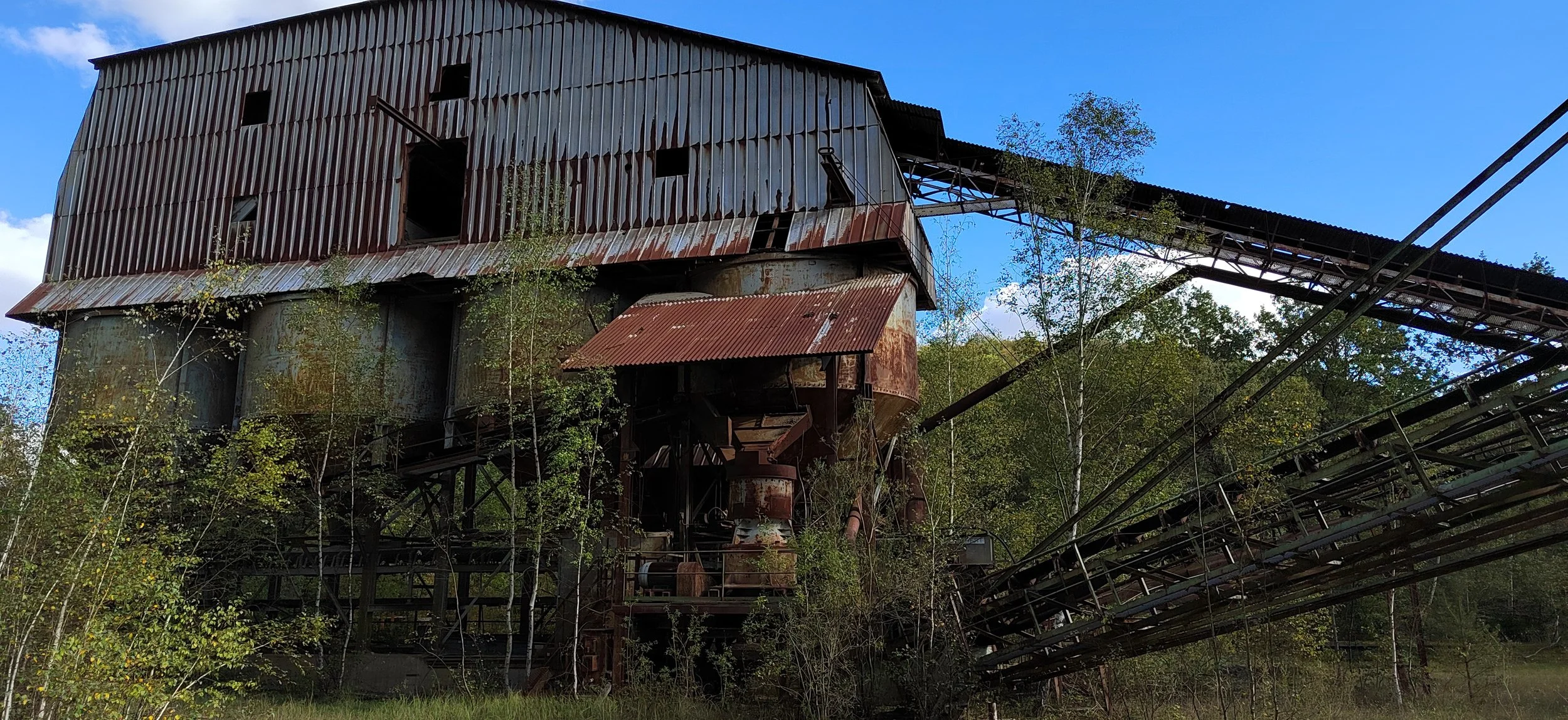 Verrostete, verlassene Industrieanlage mit einer langen, schrägen Förderbrücke und überwachsenen Pflanzen im Vordergrund, blauem Himmel im Hintergrund.