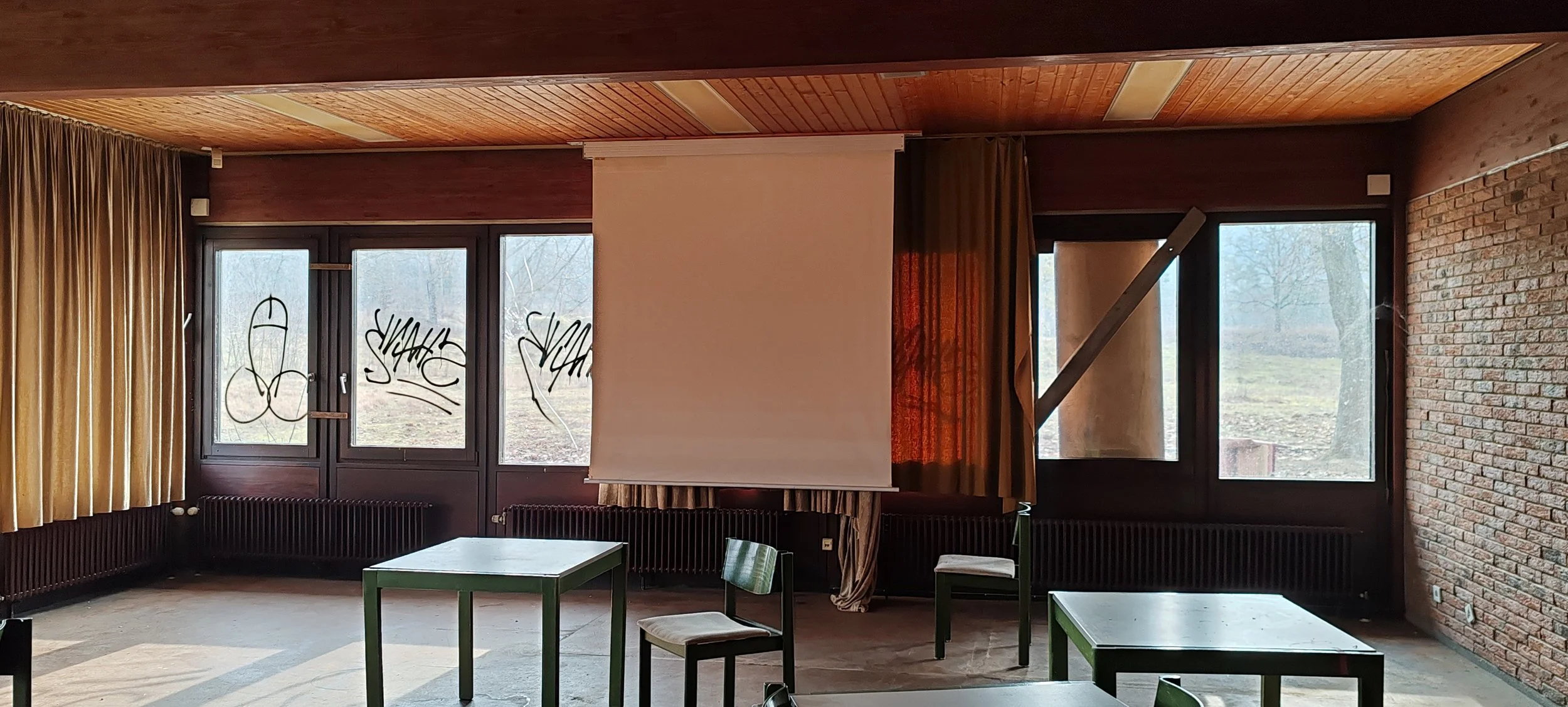 Leerer Sitzungsraum mit Tischen, Stühlen, Vorhang, Projektorleinwand, große Fenster mit Blick nach draußen, und graffiti an den Fenstern.