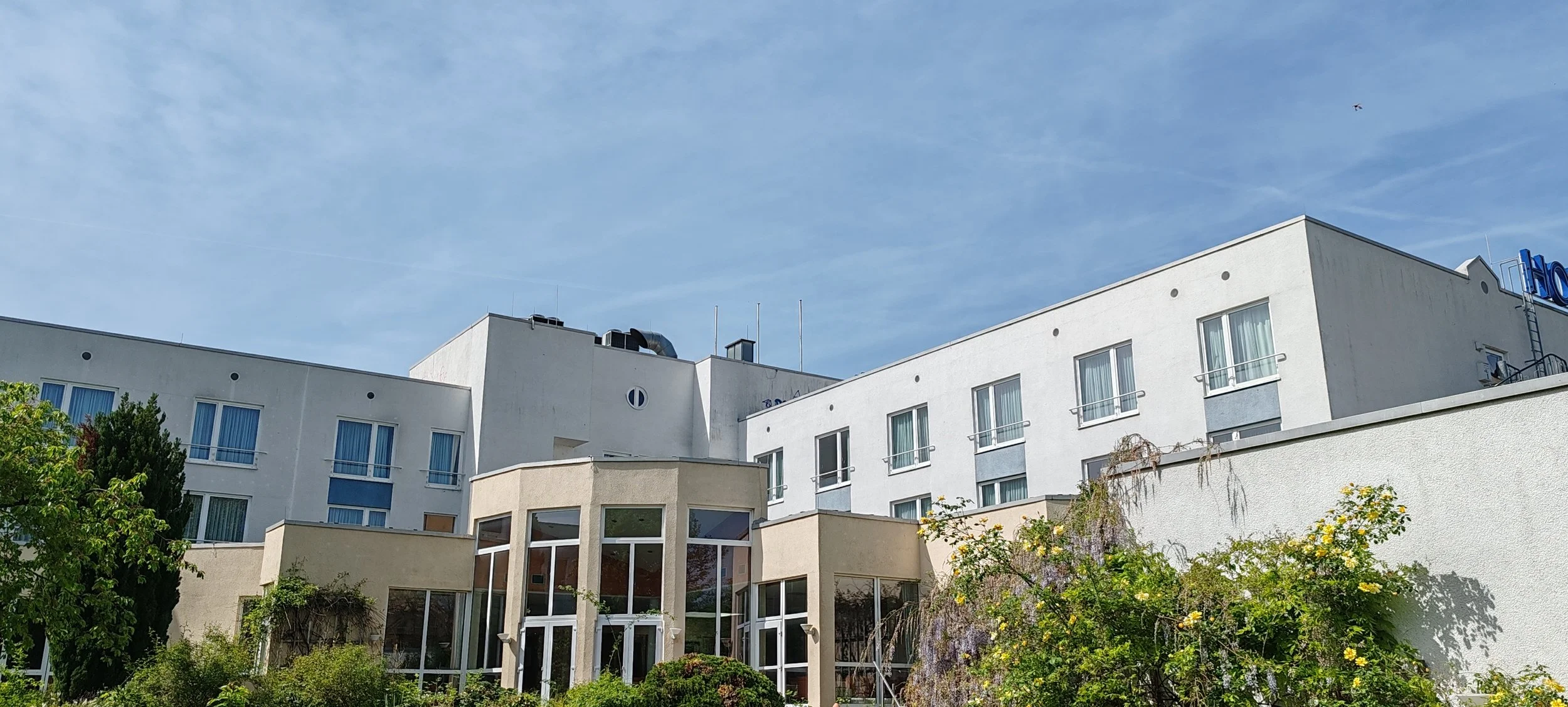 Mehrstöckiges weißes Gebäude mit großen Fenstern und Garten mit Bäumen und Sträuchern im Vordergrund, blauer Himmel mit wenigen Wolken.
