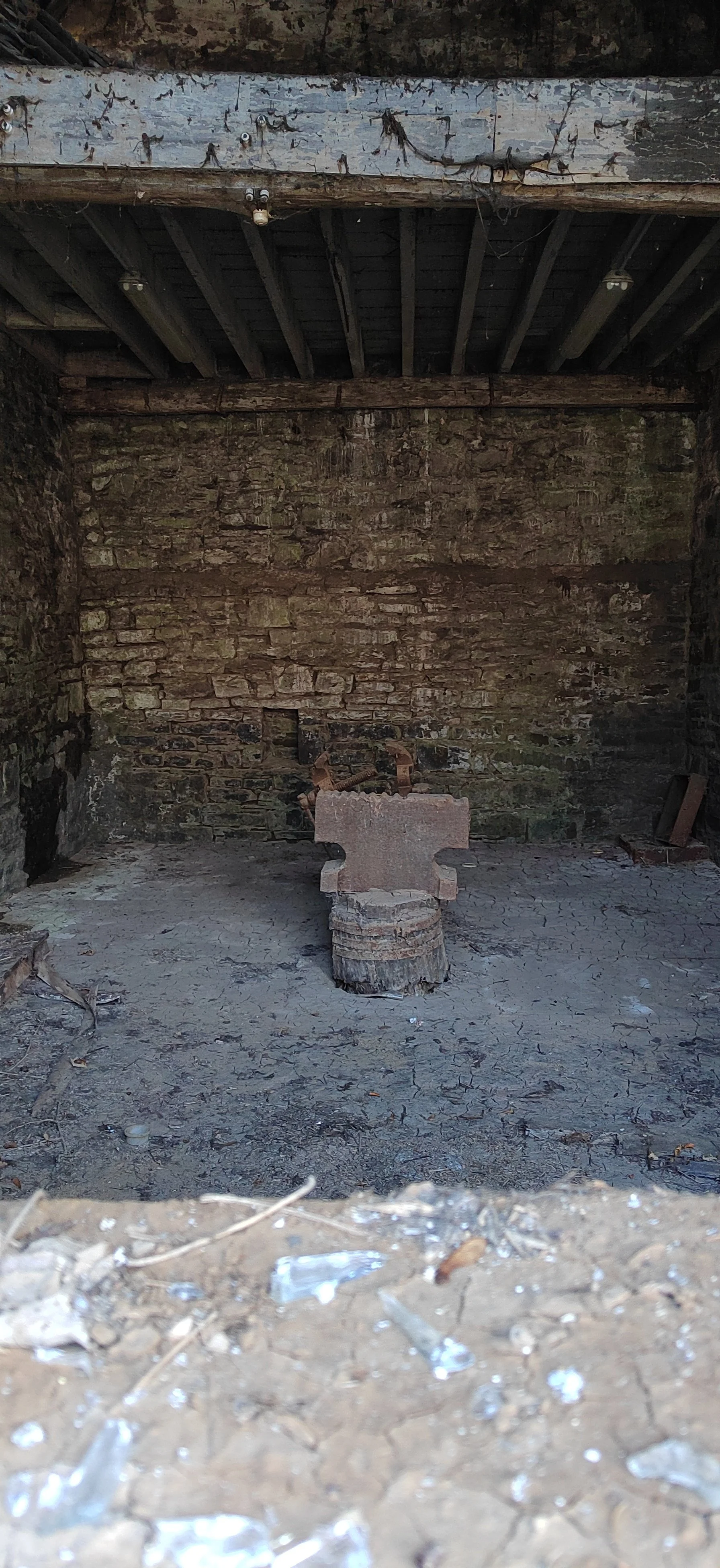 Ein verlassener Raum mit einer steinernen Kochplatte auf einem Baumstamm, kahle Wände und eine Decke mit sichtbaren Holzbalken.