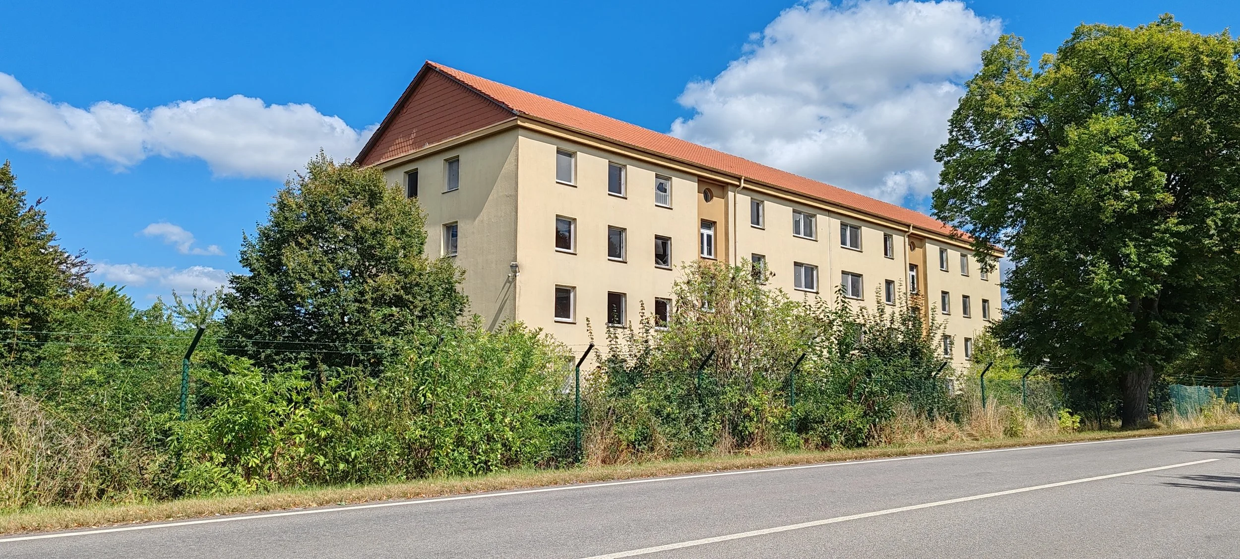 Mehrstöckiges Wohngebäude mit rotem Satteldach, umgeben von Bäumen und Sträuchern, neben einer Straße mit Doppelline in einer ländlichen Umgebung bei sonnigem Himmel.