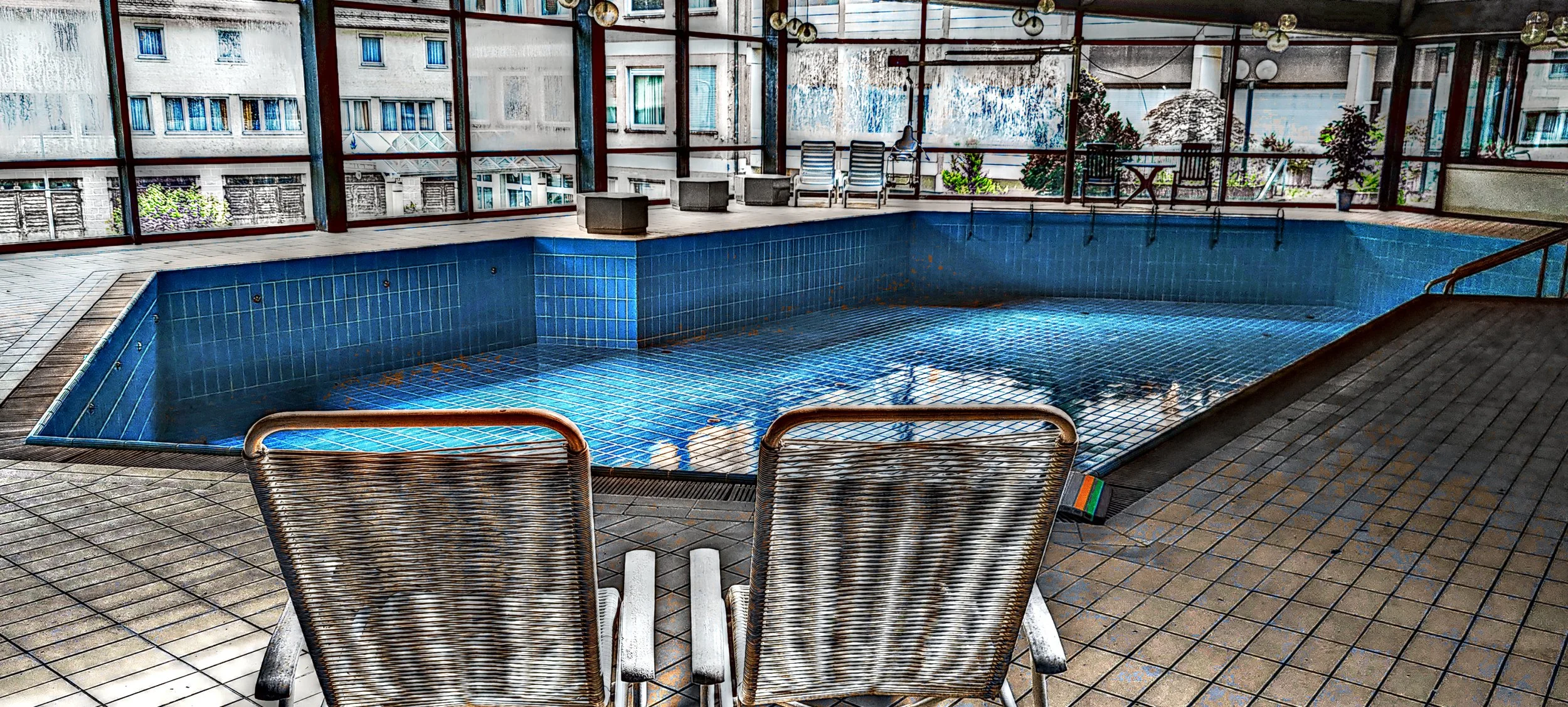 Ein verlassener, leerer Innenpool mit blauen Fliesen, umgeben von Sonnenliegen und Stühlen, mit großen Fenstern, die citysliche Gebäude im Hintergrund zeigen.