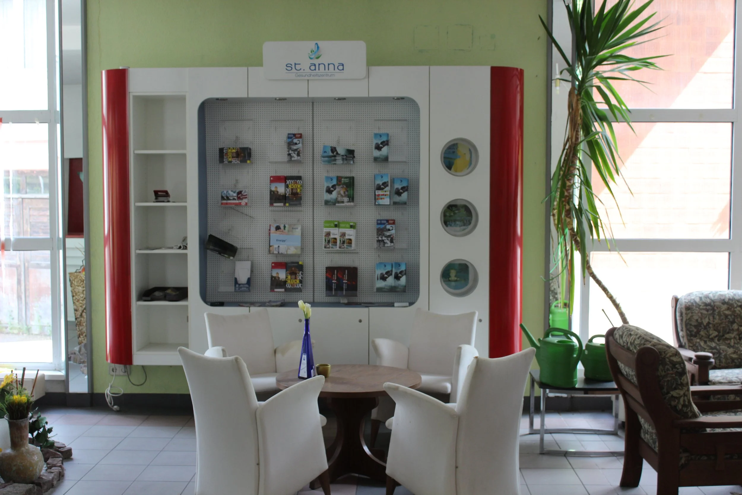 Wartebereich mit weißen Stühlen, einem runden Tisch mit Vase, grünen Wasserkrügen und einer großen Zimmerpflanze vor einem Fenster, hinter einer Vitrine mit Broschüren, im Hintergrund eine grüne Wand.