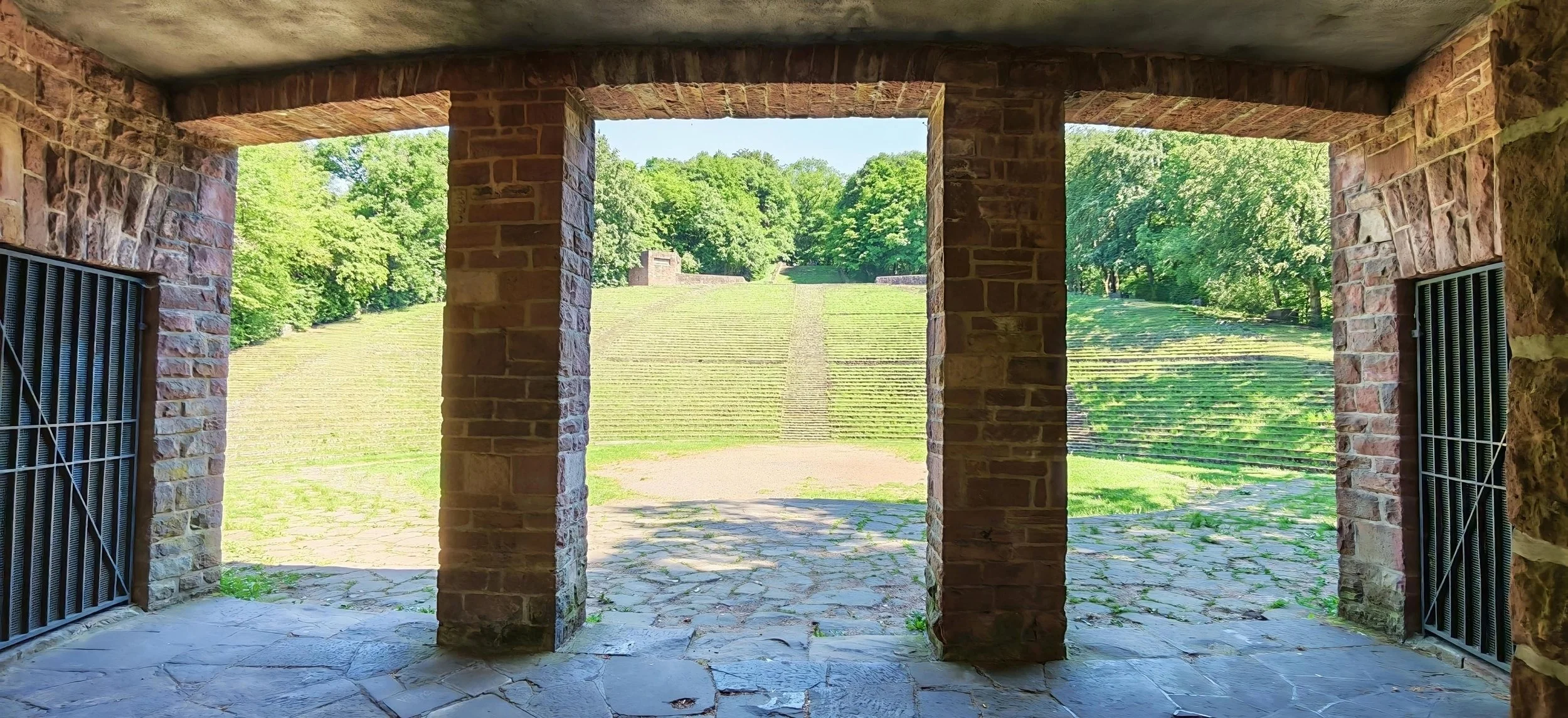 Blick durch einen offenen Torbogen auf eine terrassierte Grünfläche mit Bäumen im Hintergrund.