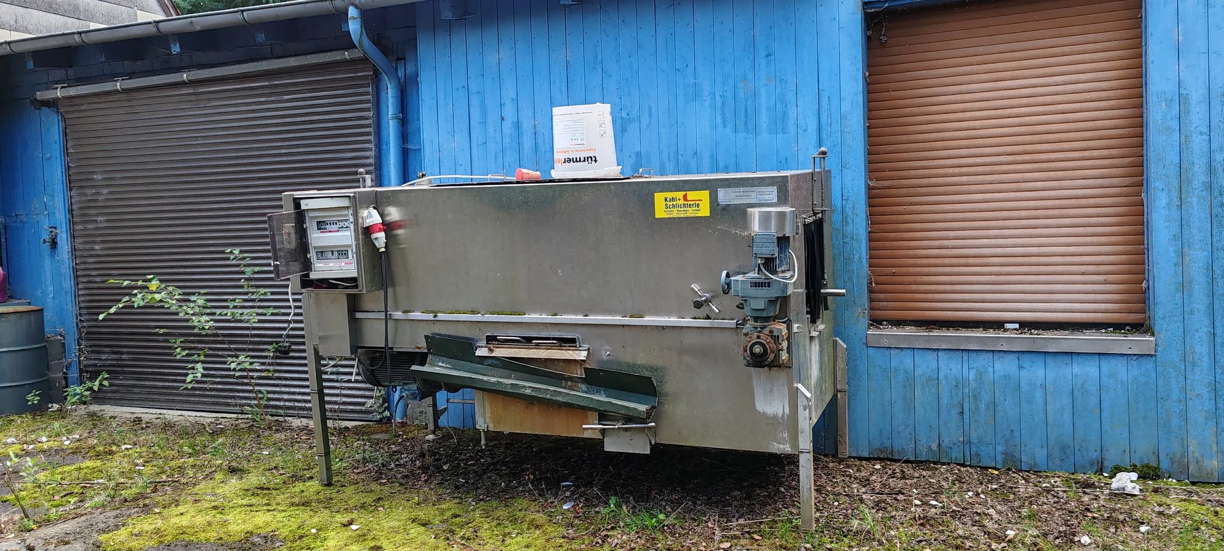 Industriegerät vor blauem Holzhaus mit geschlossener brauner Rollladenfenster, auf dem Boden mit Moos und Erde.