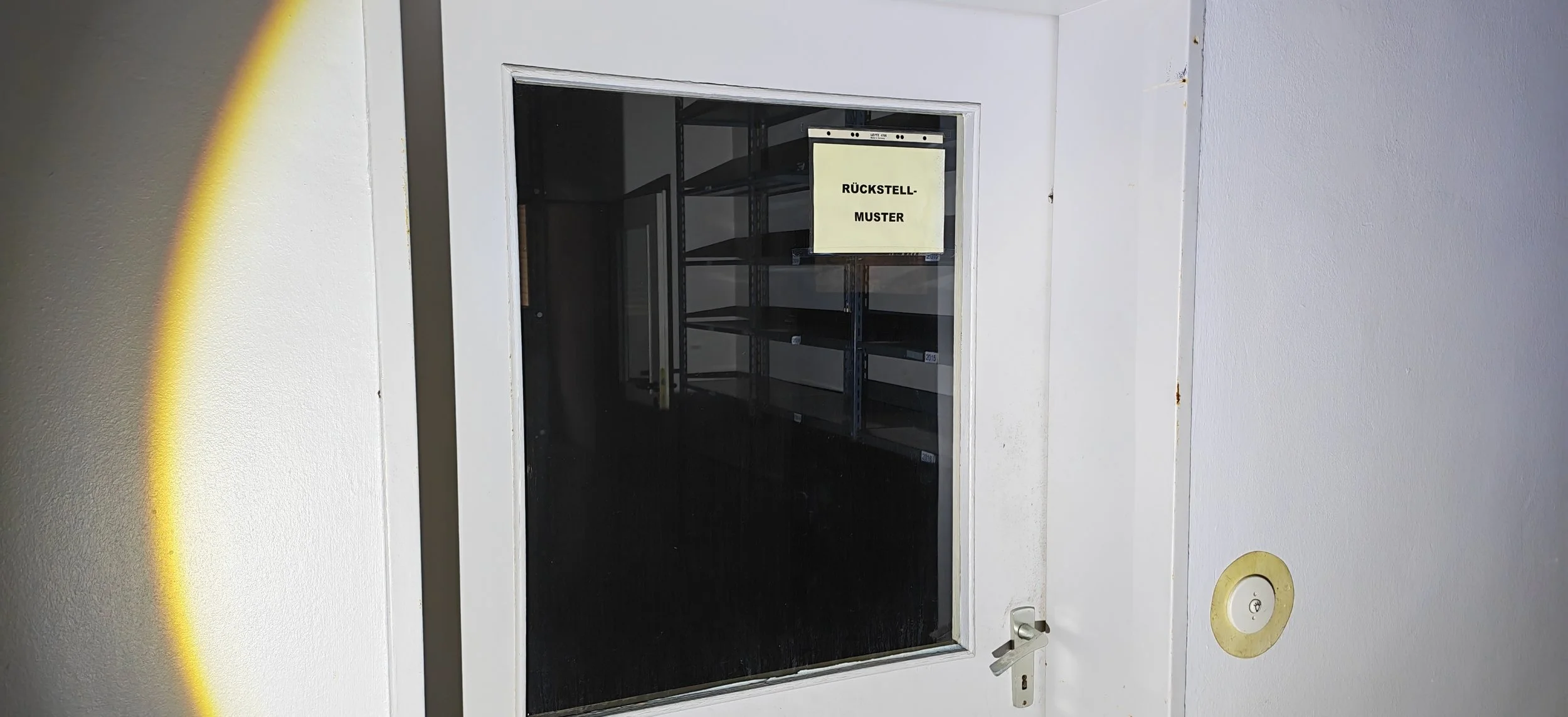 Leere Regale hinter einem Glasfenster mit einem gelben Schild, auf dem 'RÜCKSTELL-MUSTER' steht, in einem dunklen Raum, Blick durch eine geschlossene Tür