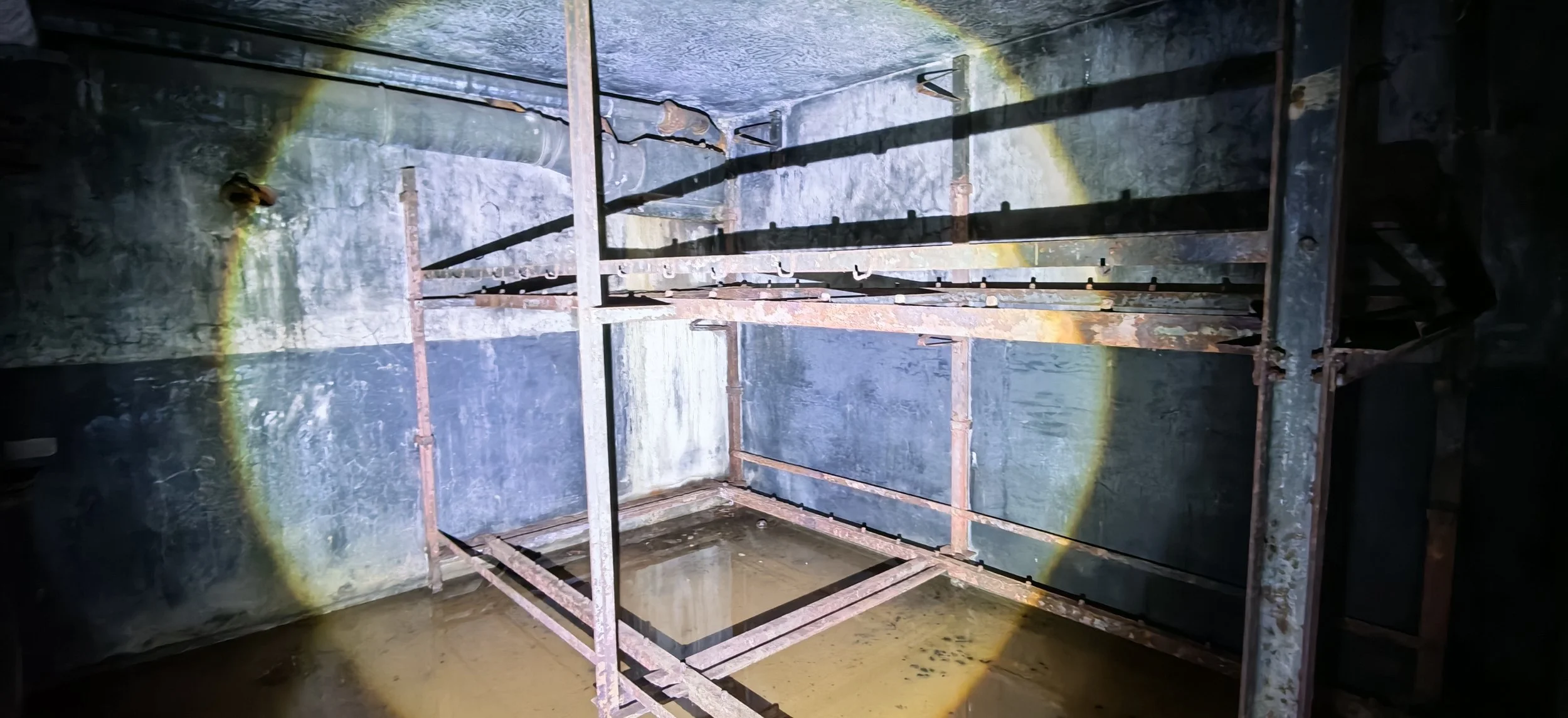 Innenansicht eines alten, verrosteten Aufzugsschachts mit Wasseransammlung am Boden, Metallrahmen und Wandflächen mit Spuren von Rost und Wasser