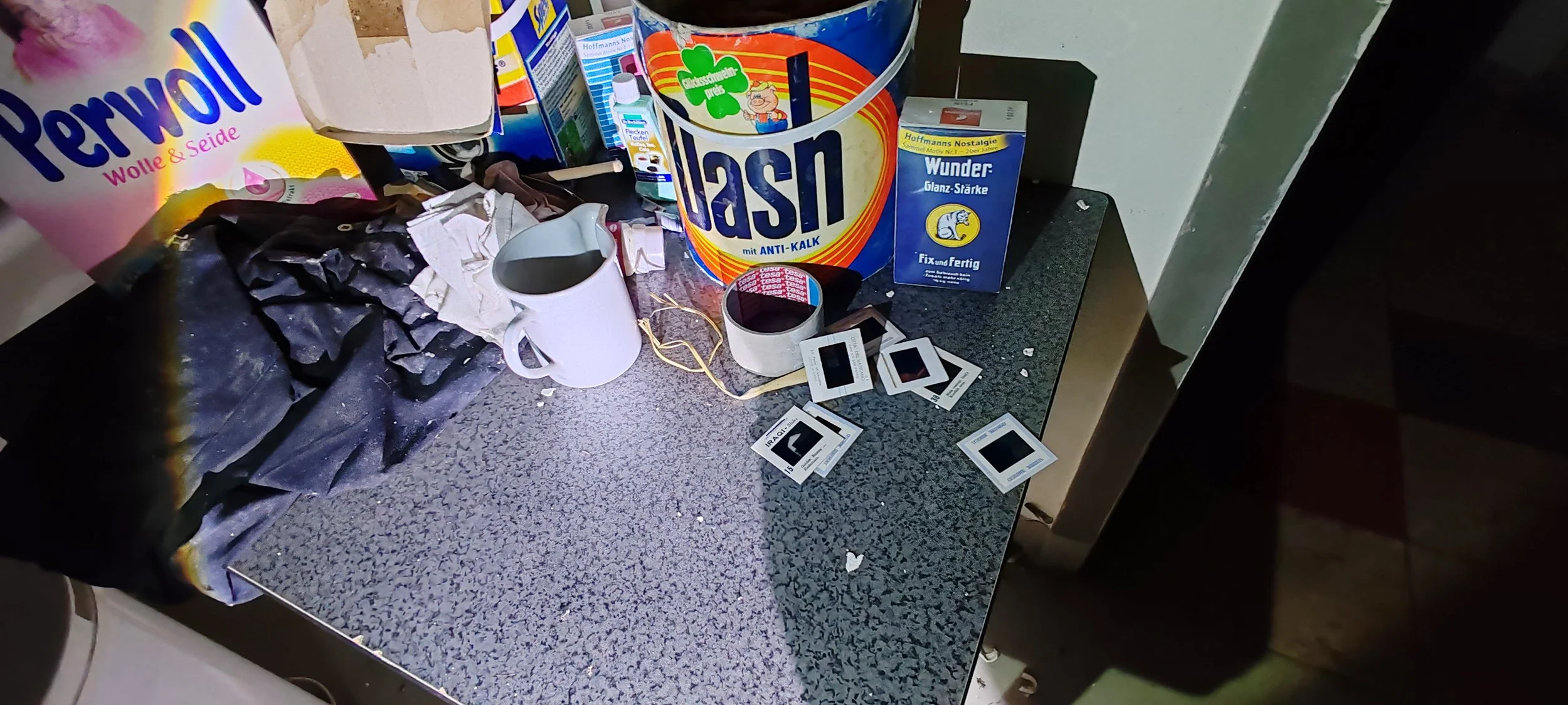 Unaufgeräumter Küchentisch mit Reinigungsmitteln, Tassen, Teelichtern, Zigarettenschachteln, Papier und Staub.