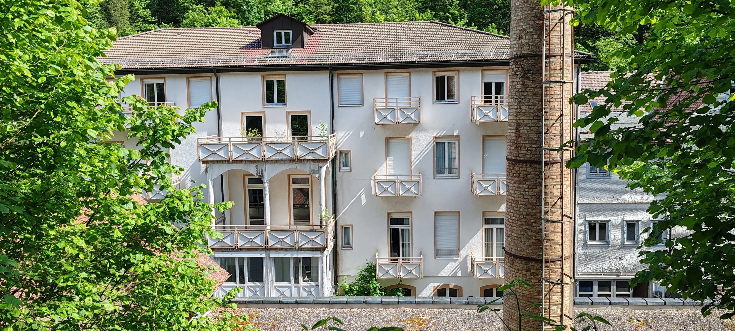 Mehrstöckiges weißes Wohngebäude mit Balkonfronten und Dachgarten, umgeben von grünen Bäumen, mit einem alten Backstein-Schornstein im Vordergrund.