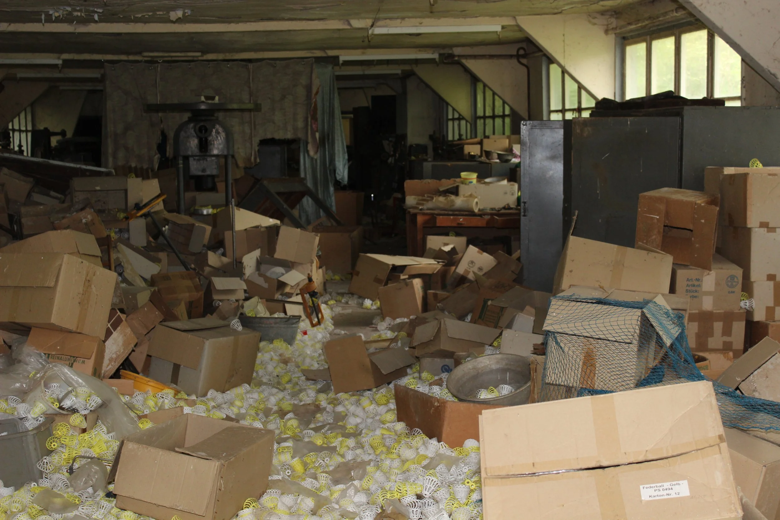Ein unordentlicher Raum voller Kartons, Plastikflaschen und Verpackungen, vermutlich eine Lagerhalle oder ein Abstellraum.