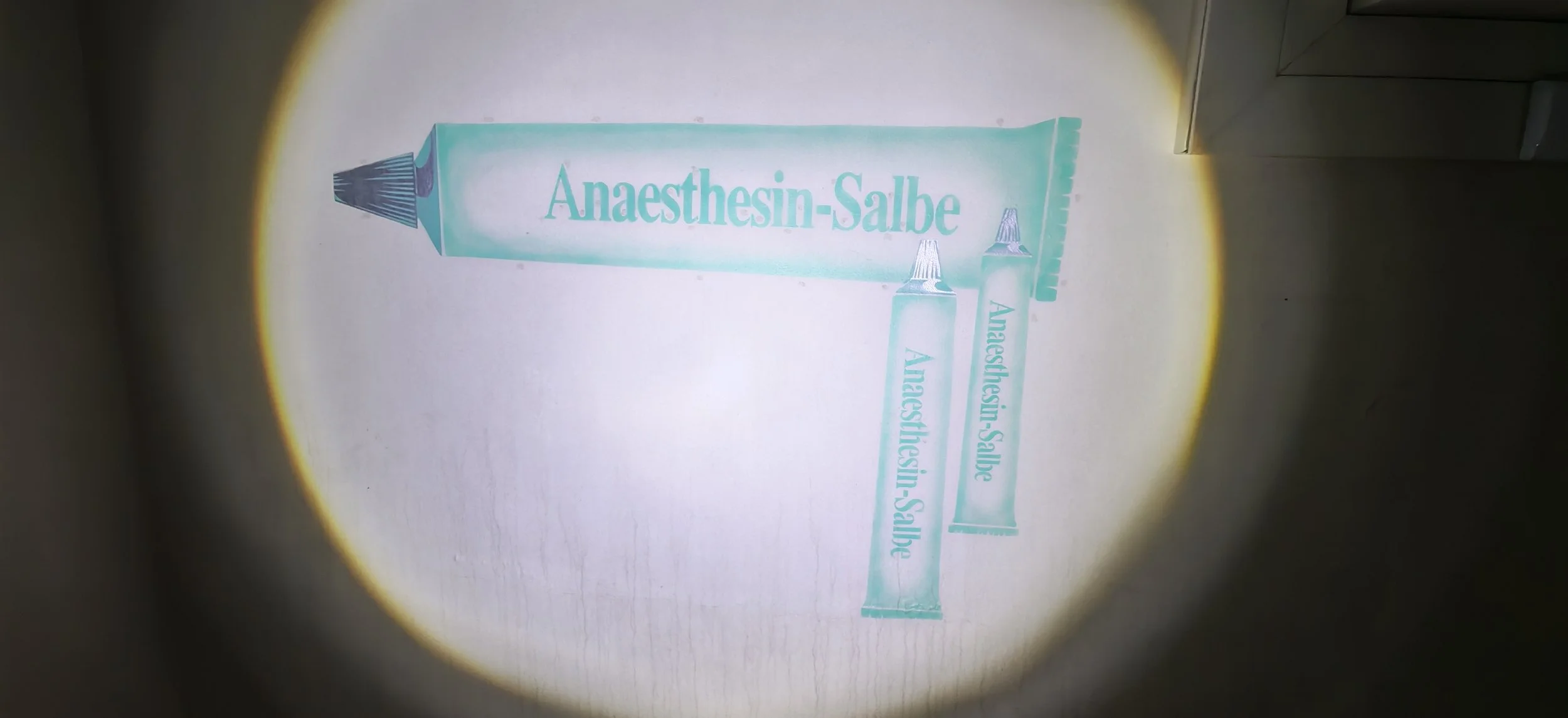 Nahaufnahme von einer Tube mit der Aufschrift 'Anaesthesin-Salbe' auf einer hellen Oberfläche, beleuchtet von einem runden Lichtfleck, vermutlich eine medizinische Salbe. Zwei kleinere Tuben mit gleicher Beschriftung liegen daneben.
