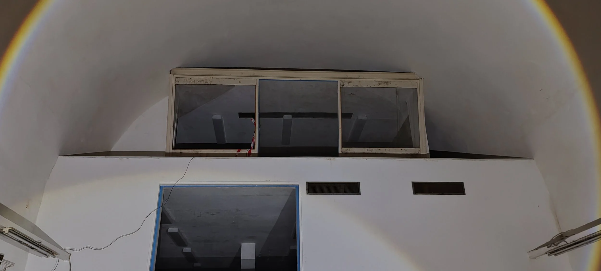 Innenaufnahme eines Raums mit weißen Wänden, einem großen Fenster an der Decke und kleinen Lüftungsschlitzen an der Wand. Es gibt eine sichtbare Leitung, die an der Wand befestigt ist.