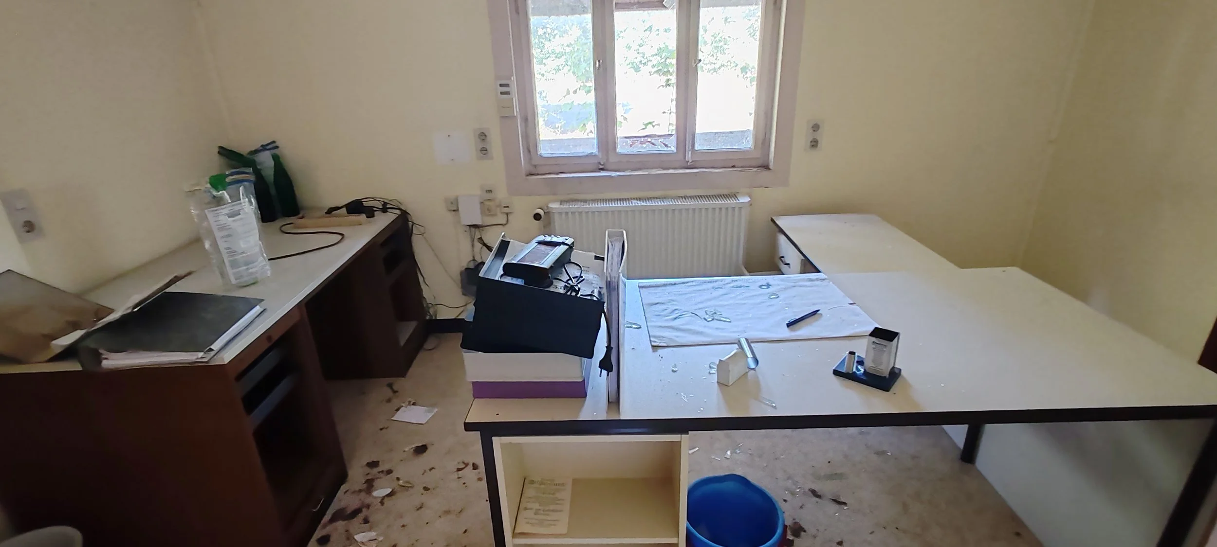 Ein unordentlicher Raum mit einem Schreibtisch, einigen Papieren und Gegenständen, die auf dem Tisch liegen, sowie einem Fenster im Hintergrund und einem beschädigten Boden.