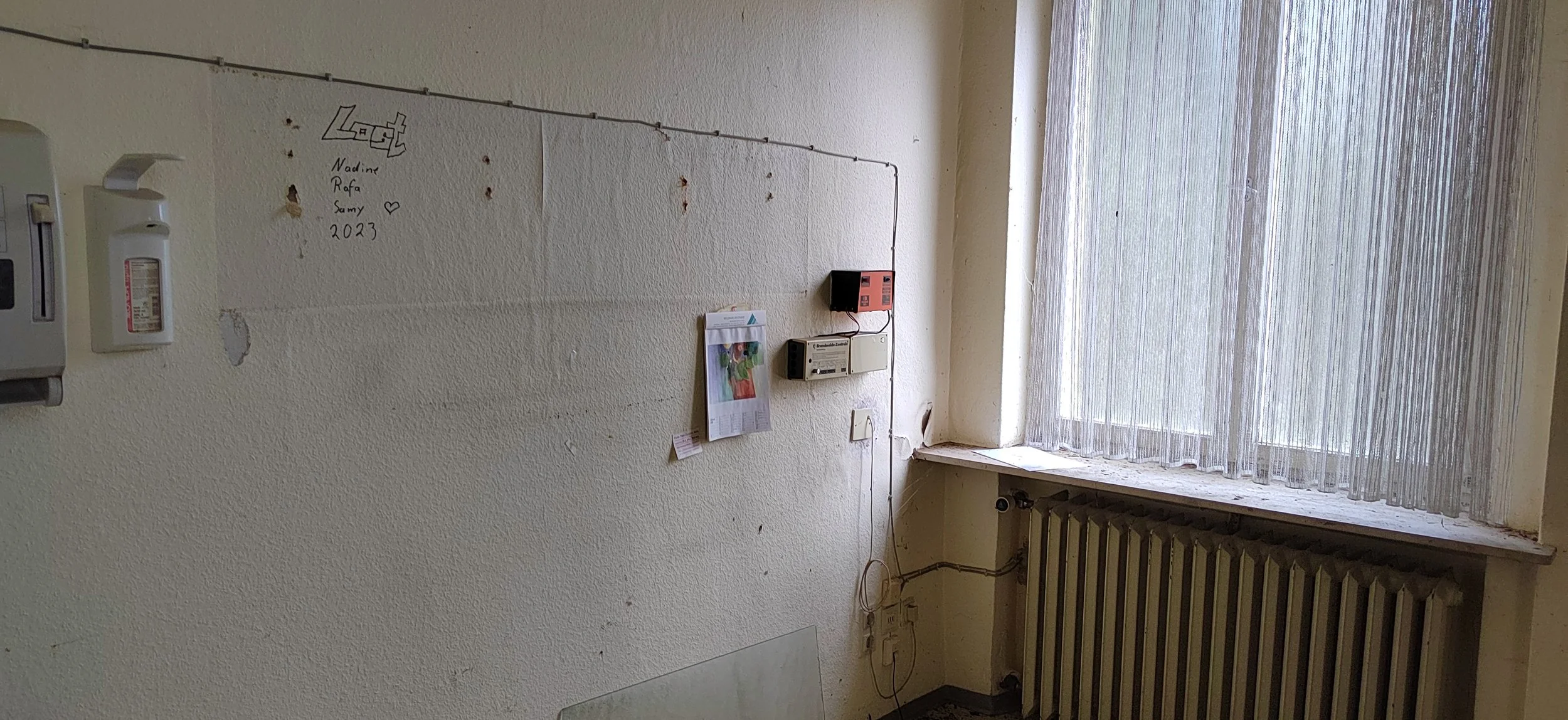 Ein alter Raum mit einer verputzten Wand, einem großen Fenster mit halbtransparenter Gardine, einem Heizkörper und mehreren Gegenständen an der Wand, darunter eine Notrufbox, ein Notizblock und ein kleines schwarzes Gerät.