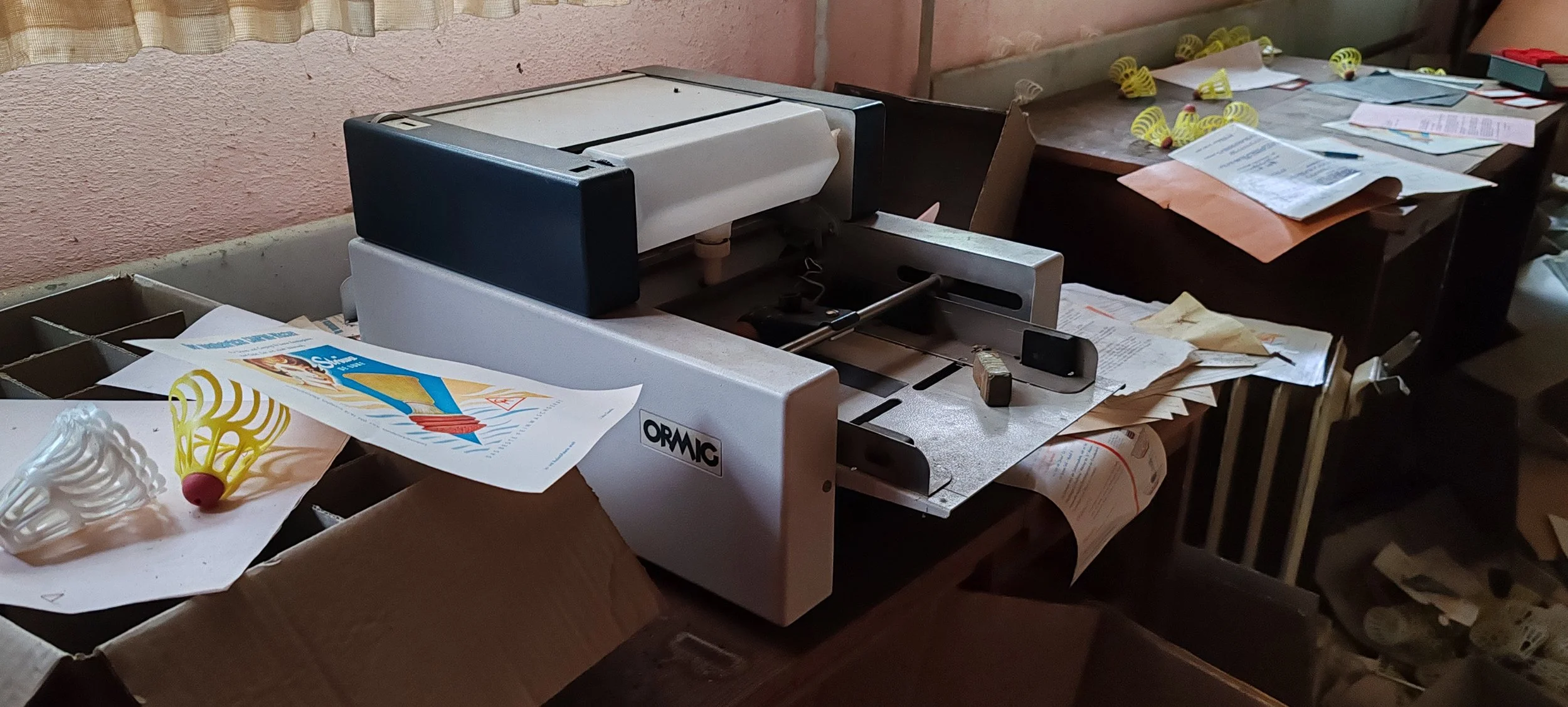 Büro- oder Bastelraum mit einem Schneidegerät, Papieren, und Bastelmaterialien. Bunte Plastik- und Papierobjekte liegen auf Tischen und Papierstapeln.