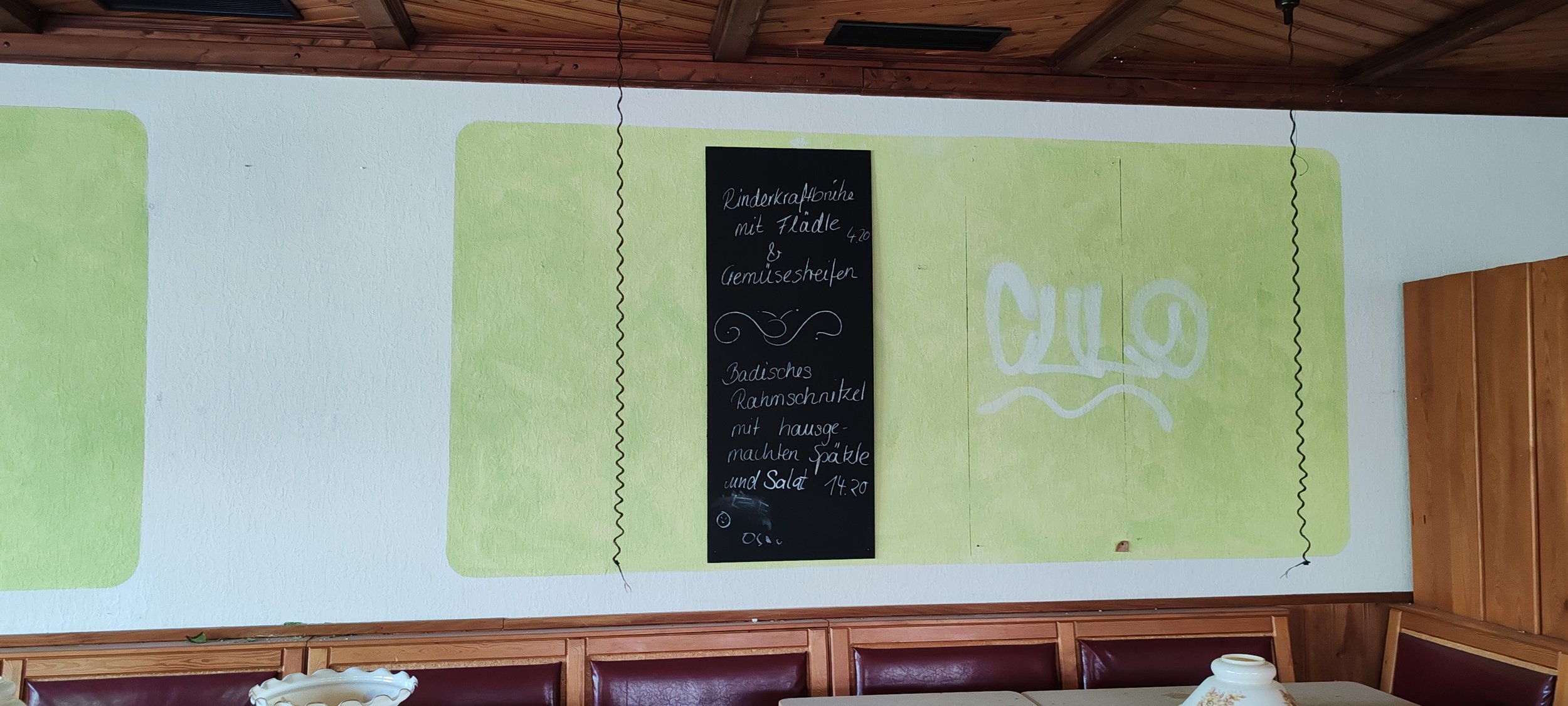 Eine grüne Wand mit einem schwarzen Speiseplan. Der Plan bietet Rinderkraftbrühe mit Flädle und Gemüsescheiben sowie Badisches Rahmschnitzel mit hausgemachten Spätzle und Salat an.