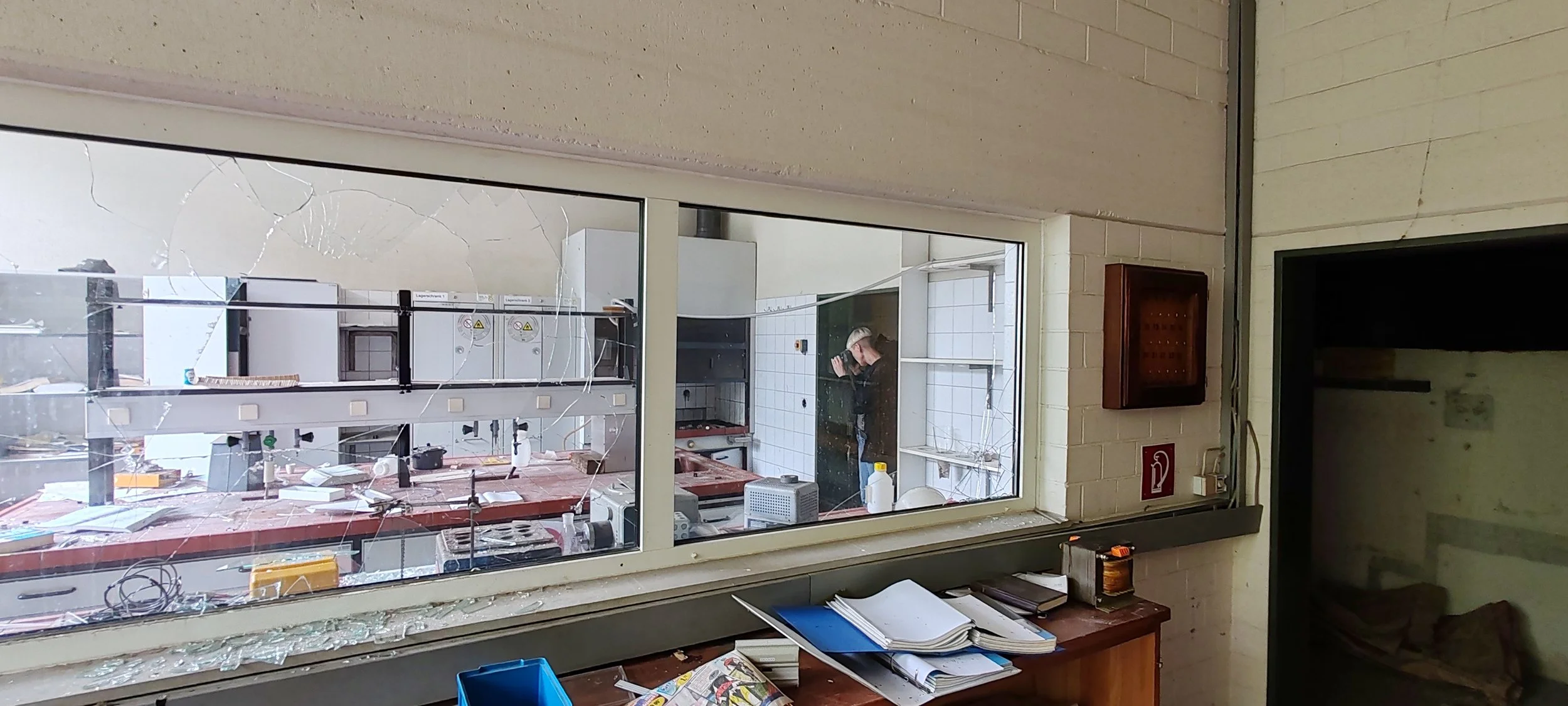 Blick durch ein zerbrochenes Fenster in einen Raum, in dem Schriftzüge auf der Scheibe Dekor- und Laborgeräte auf einem Tisch sowie eine Person bei der Arbeit sichtbar sind.