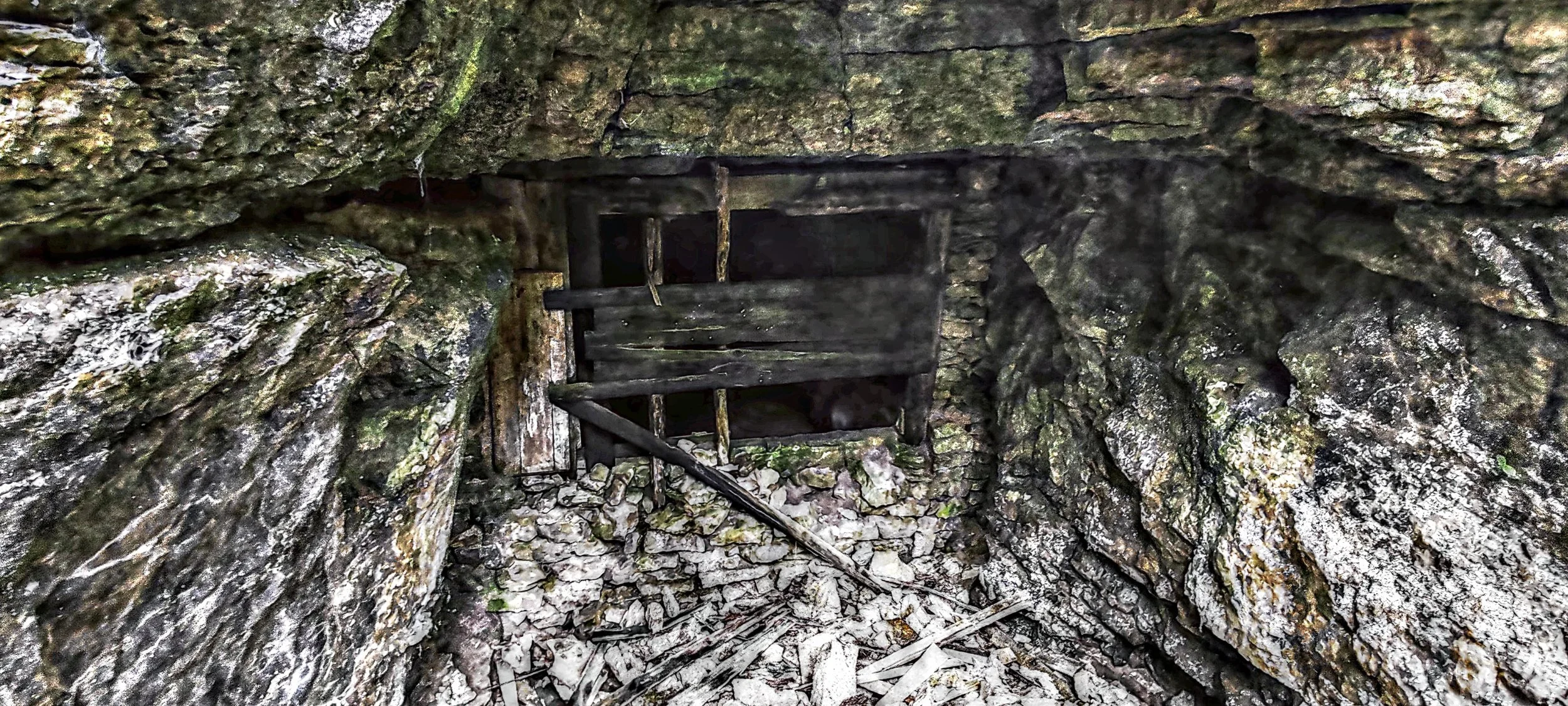 Verfallene Holztreppe in einer Höhle mit rauen, mossbedeckten Felswänden und aufgelöstem Boden aus Steinen und Holzsplitt.