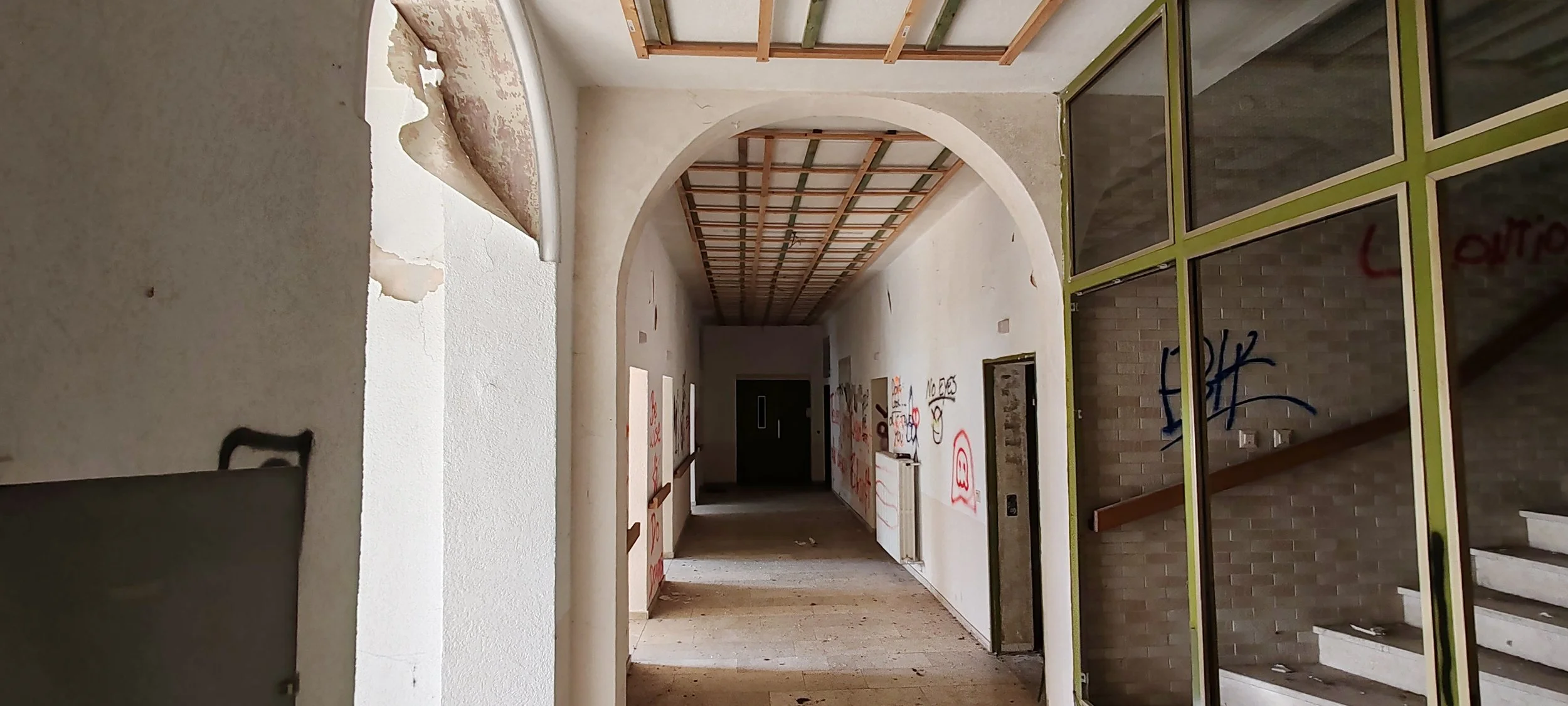 Innenansicht eines alten, ungepflegten Gebäudeteils mit grauer Wand und Stufen, Graffiti an den Wänden und einer bröckelnden Decke mit Holzrahmen.