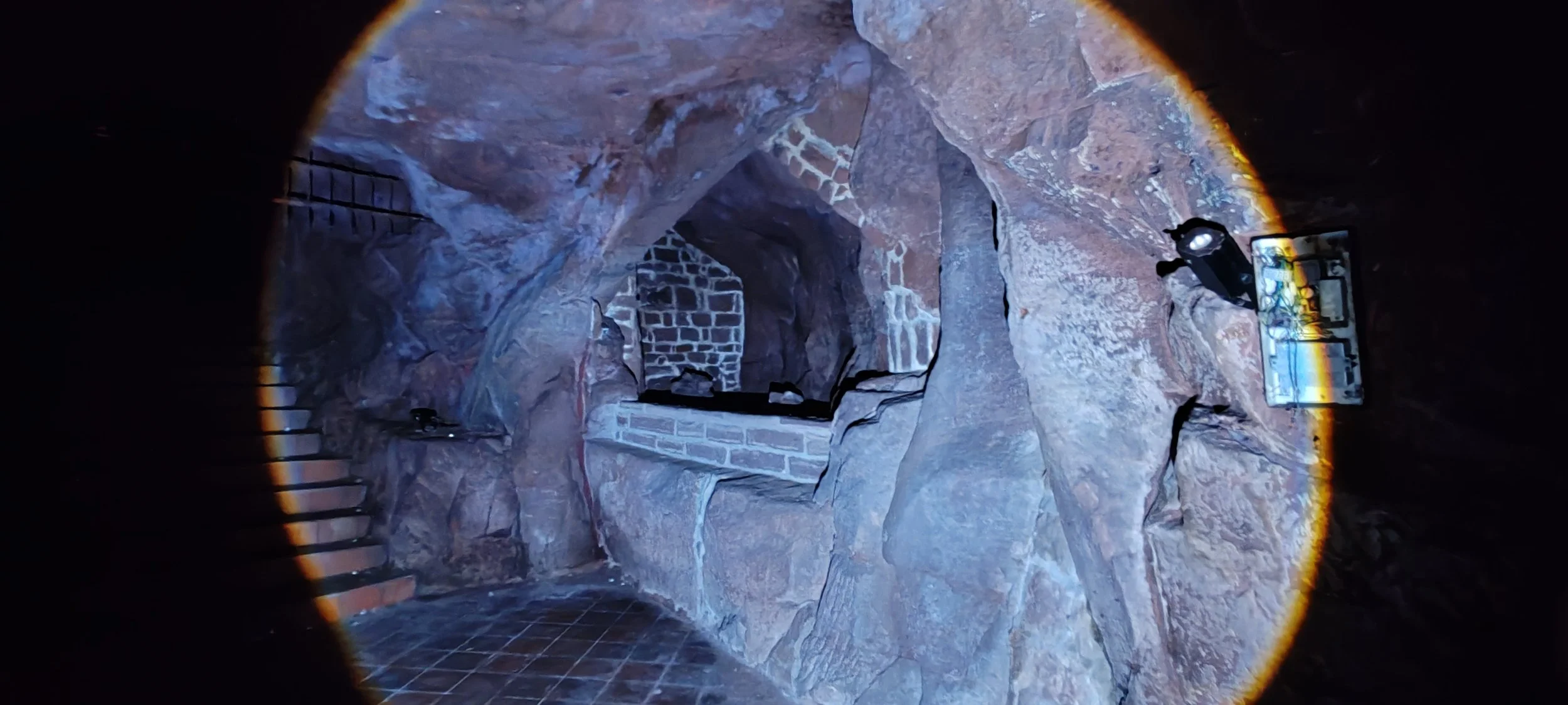 Blick in eine kleine Höhle mit Stein- und Ziegelmauern, deutlich sichtbare Treppen auf der linken Seite, beleuchtet mit einer Taschenlampe.