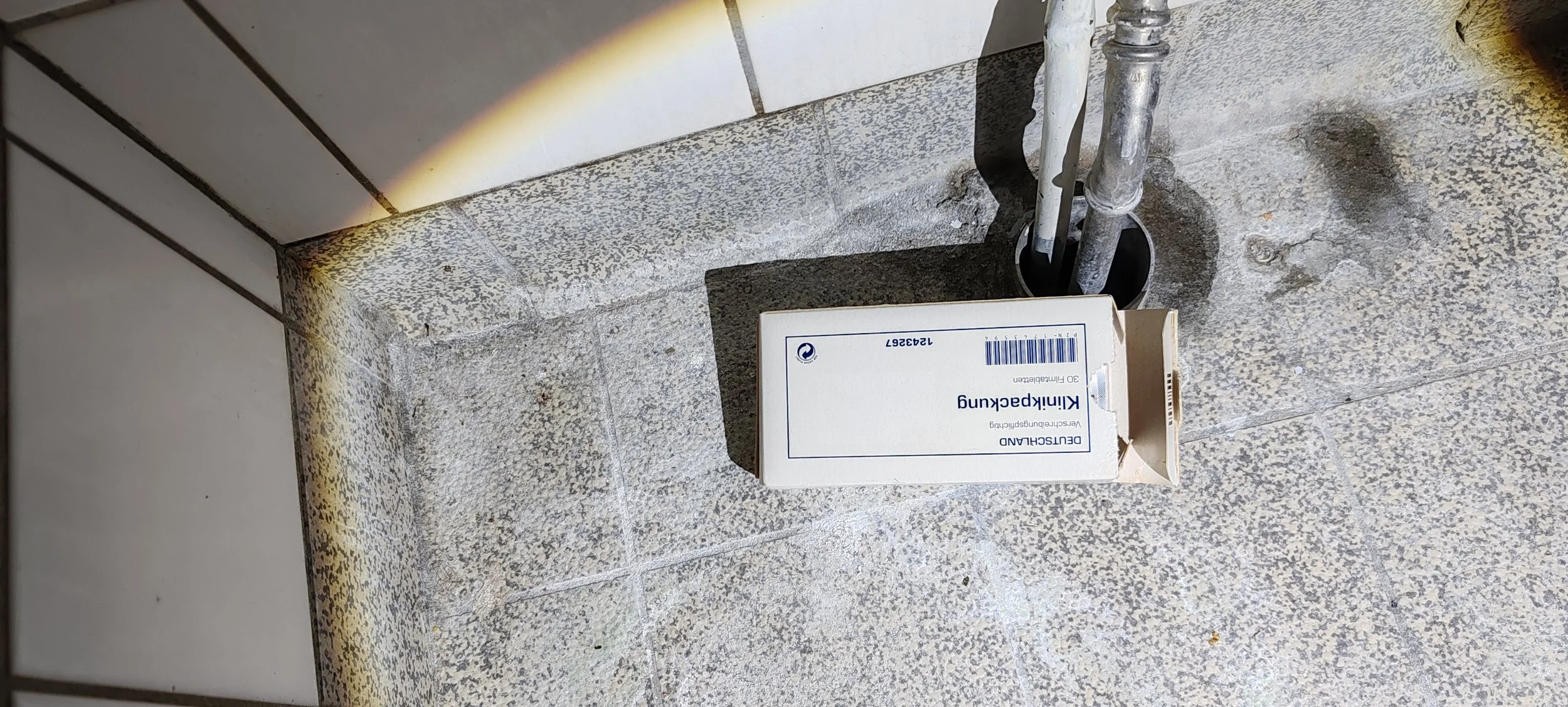Eine weiße Medizinbox mit blauer Aufschrift steht auf einem Steinboden, neben einem Metallrohr in einer Ecke.