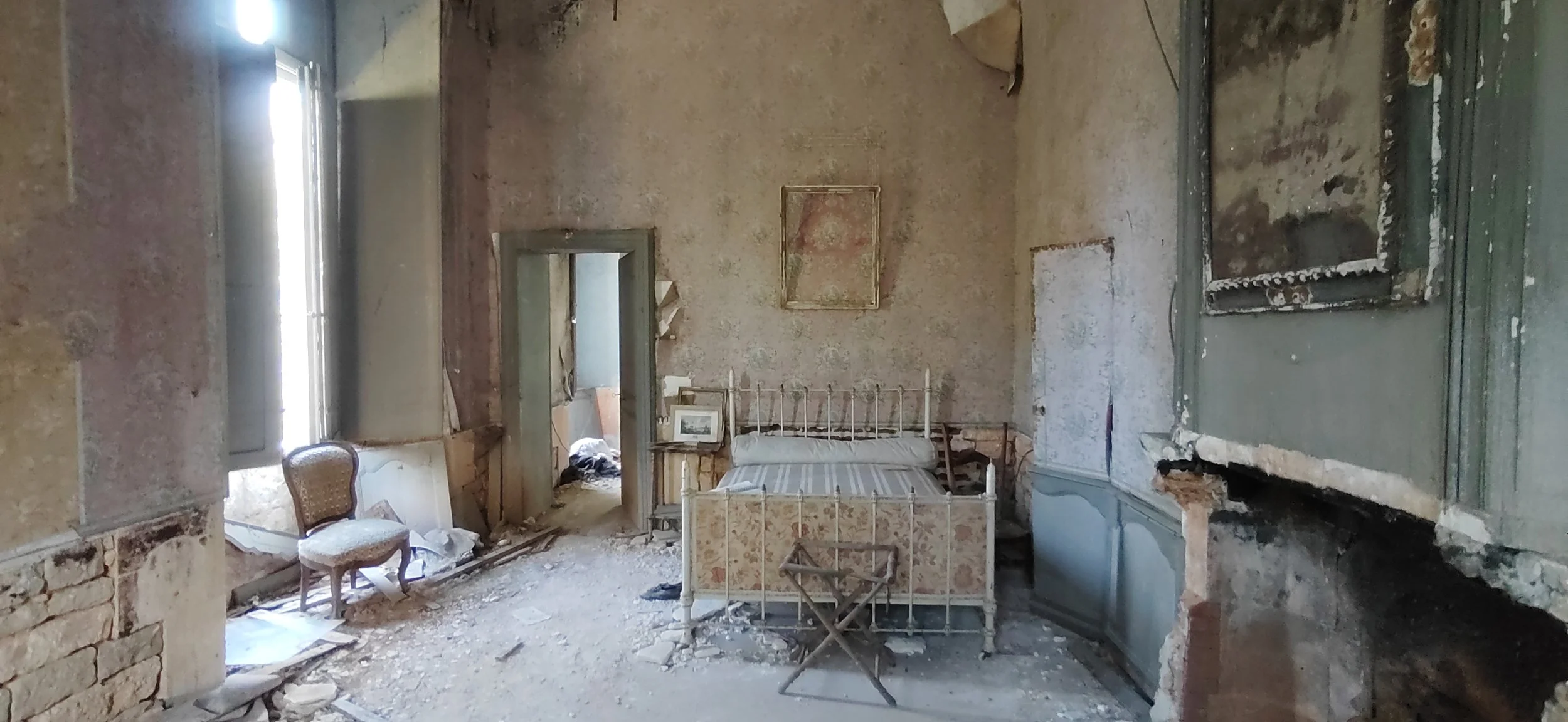 Ein altes, verfallenes Schlafzimmer mit einem verzierten Bett, einem Stuhl, renovierungsbedürftigen Wänden, morschem Boden und beschädigten Fenstern.