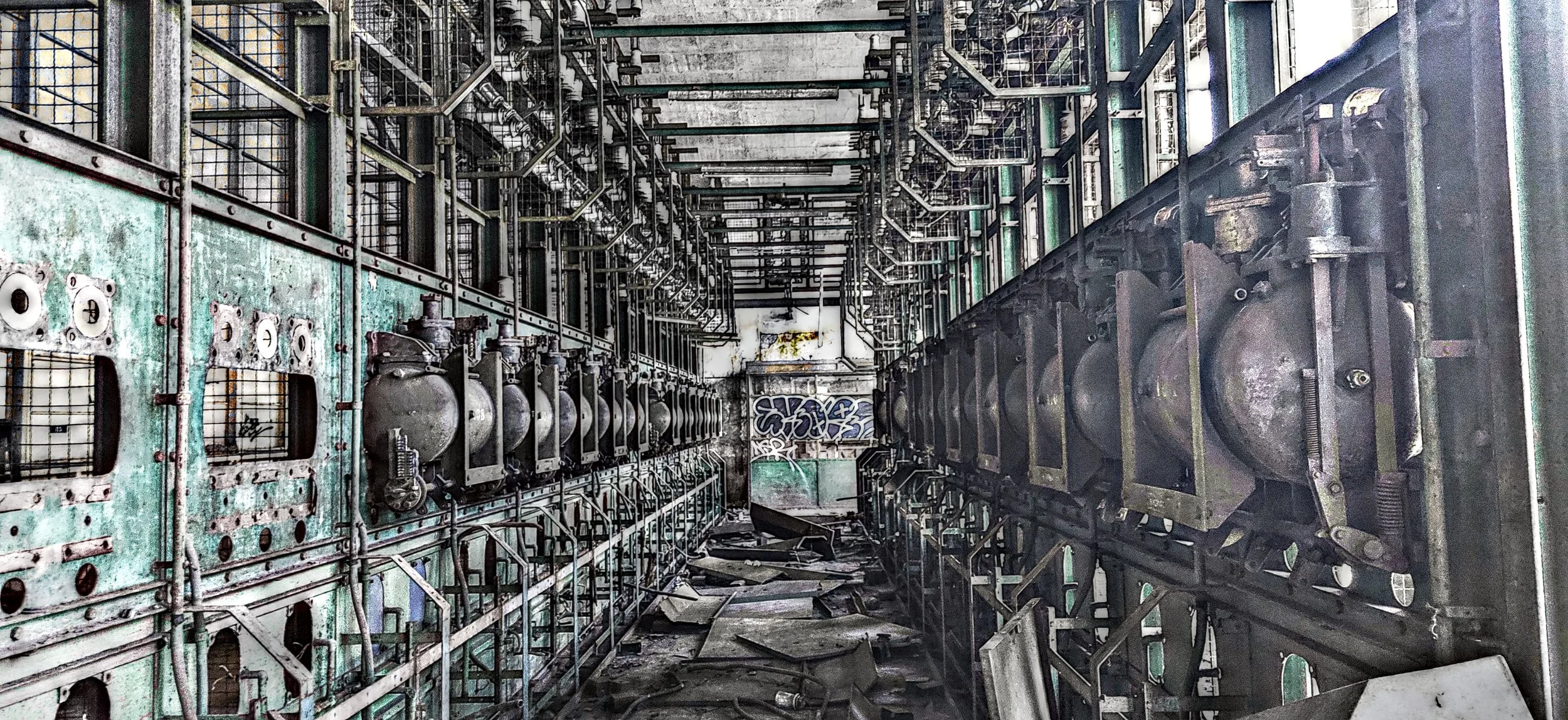 Verlassene Industriehalle mit verrosteten Maschinen, zerbrochenem Boden und Graffiti an der Wand im Hintergrund.