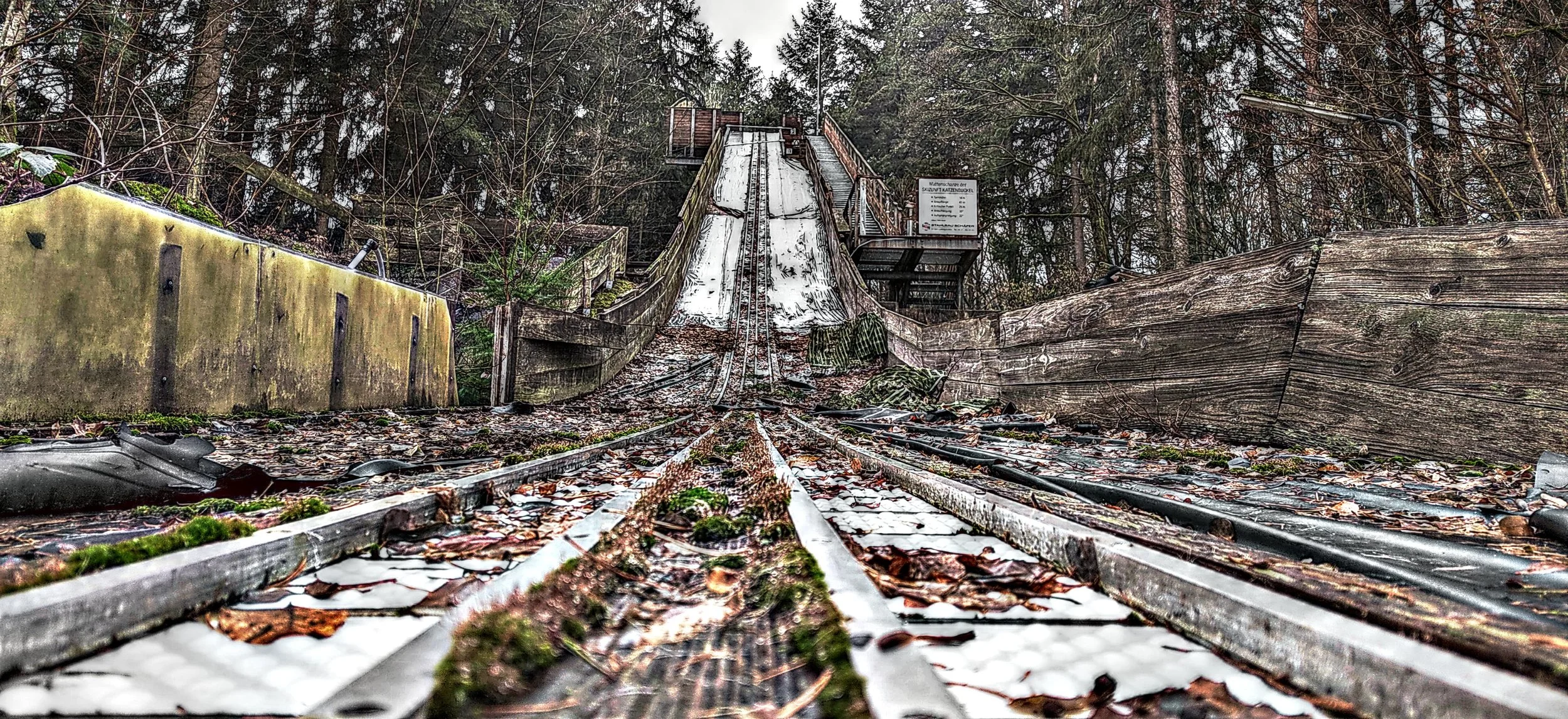 Eine verlassene Rodelbahn im Wald, mit schneebedeckten Schienen und umgestürzten Bäumen.