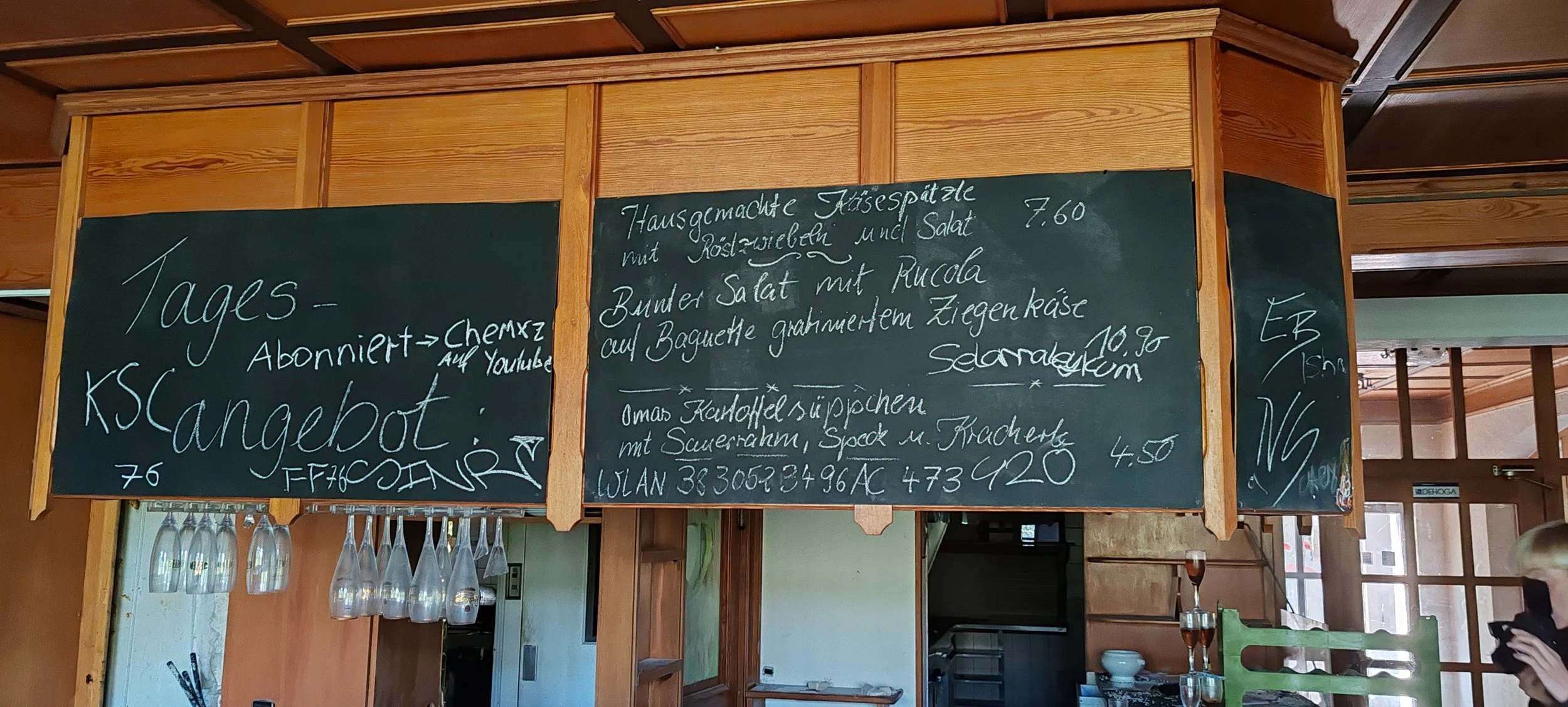 Schwarzes Wandtafel mit handgeschriebenen Angeboten und Informationen in einer Gaststube, Holzwände und Gläser hängen darunter.