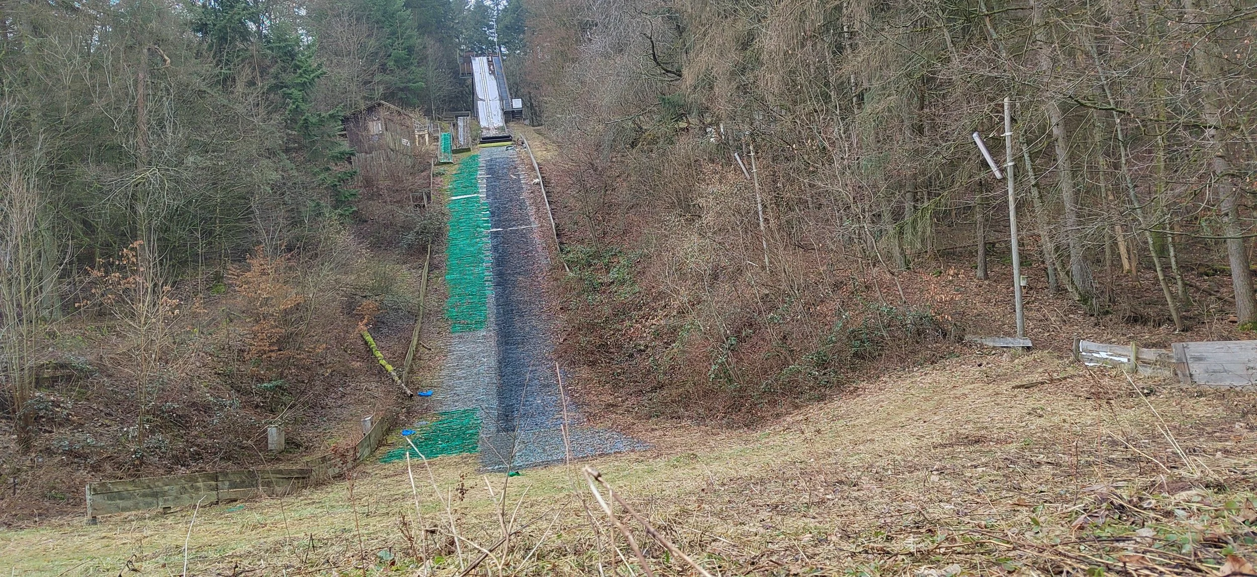 Abgesperrter Skihang im Wald, mit Schutznetz und Absperrungen am unteren Ende, sowie umgestürzten Straßenlampen und einer Holzbarriere im Vordergrund.