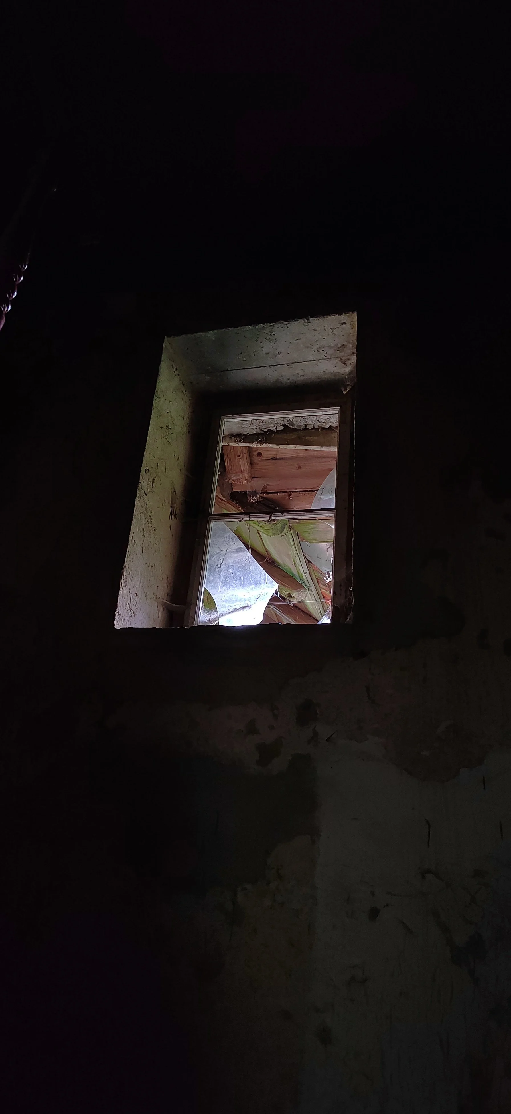 Ein kleines Fenster in einer dunklen, verfallenen Wand mit zerbrochenem Glas und sichtbarem Holz im Außenbereich.
