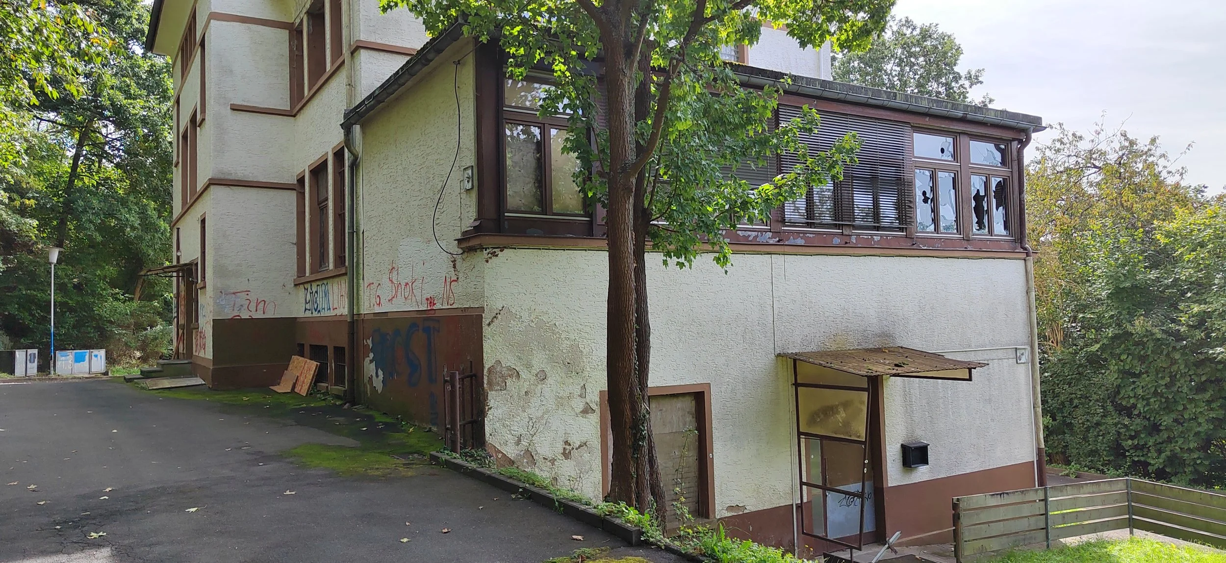 Ein älteres Mehrfamilienhaus mit teilweise veralterter Fassade, bunte Graffiti an der Wand, umgeben von Grünflächen und Bäumen, bei bewölktem Himmel.
