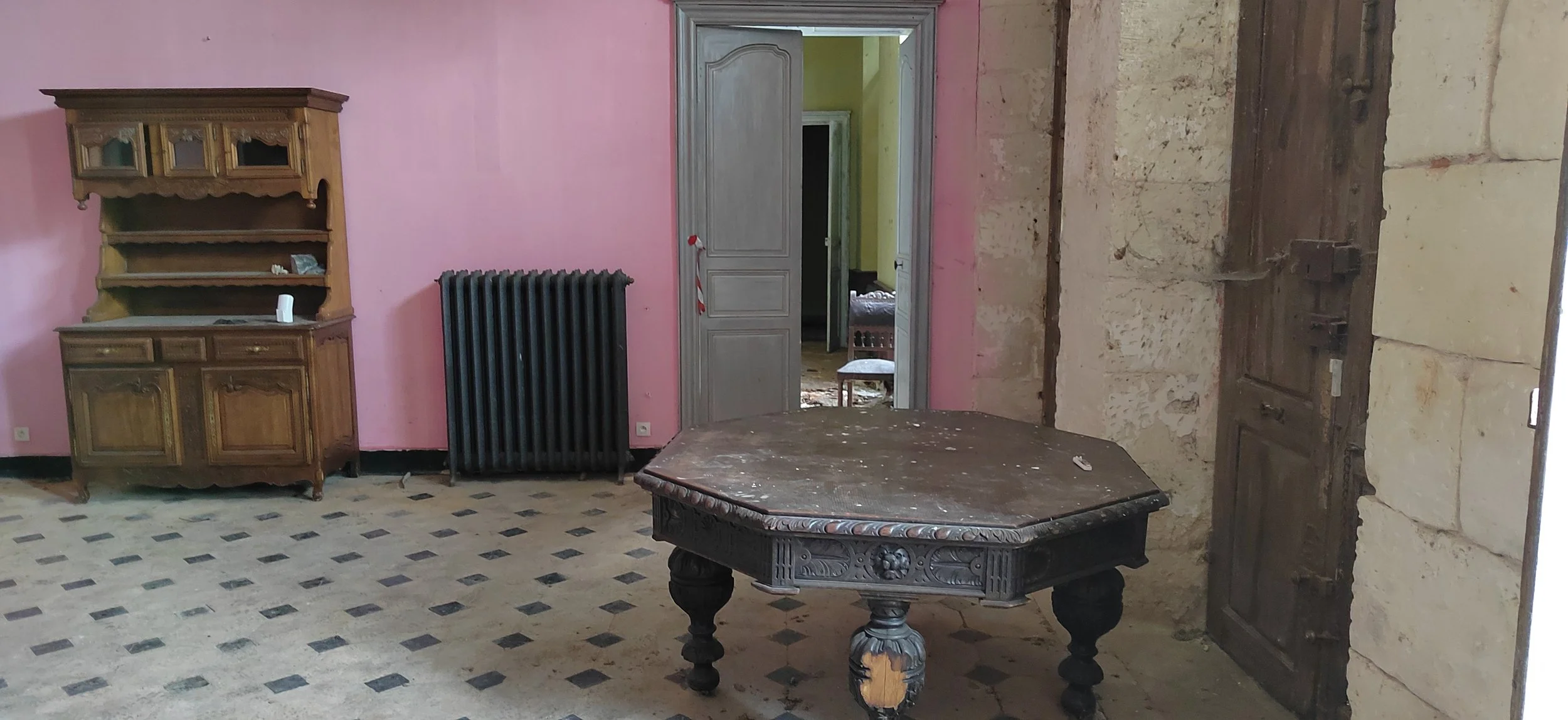 Ein altes, staubiges Wohnzimmer mit bunten Wänden, einem antiken Holzregal, einem schwarzen Heizkörper, einem großen Holztisch und einem Holzportal zu einem anderen Raum.