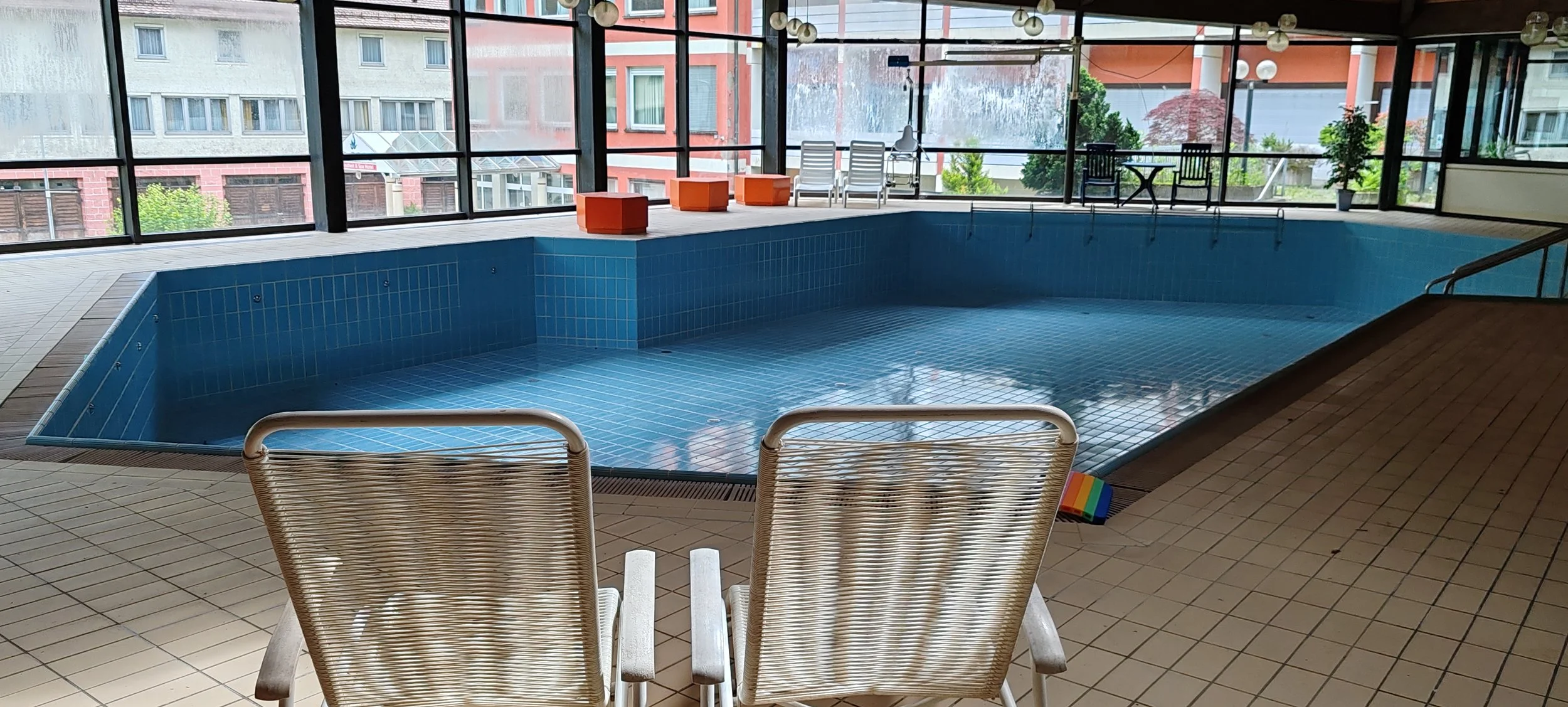 Indoor Schwimmbad mit blauer Fliesen, Sonnenliegen, Stühlen und Tischen, große Fenster mit Blick auf Nachbargebäude und Balkon, Rutschmatte am Poolrand.