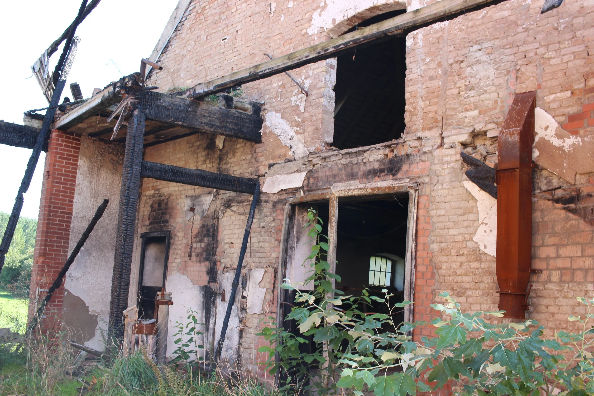 Verbrannte und beschädigte Backsteinwand eines Hauses mit schwarzen Brandspuren, aufgerissenen Fenstern und verbrannten Holzstrukturen. Pflanzen wachsen vor der Hausfassade.