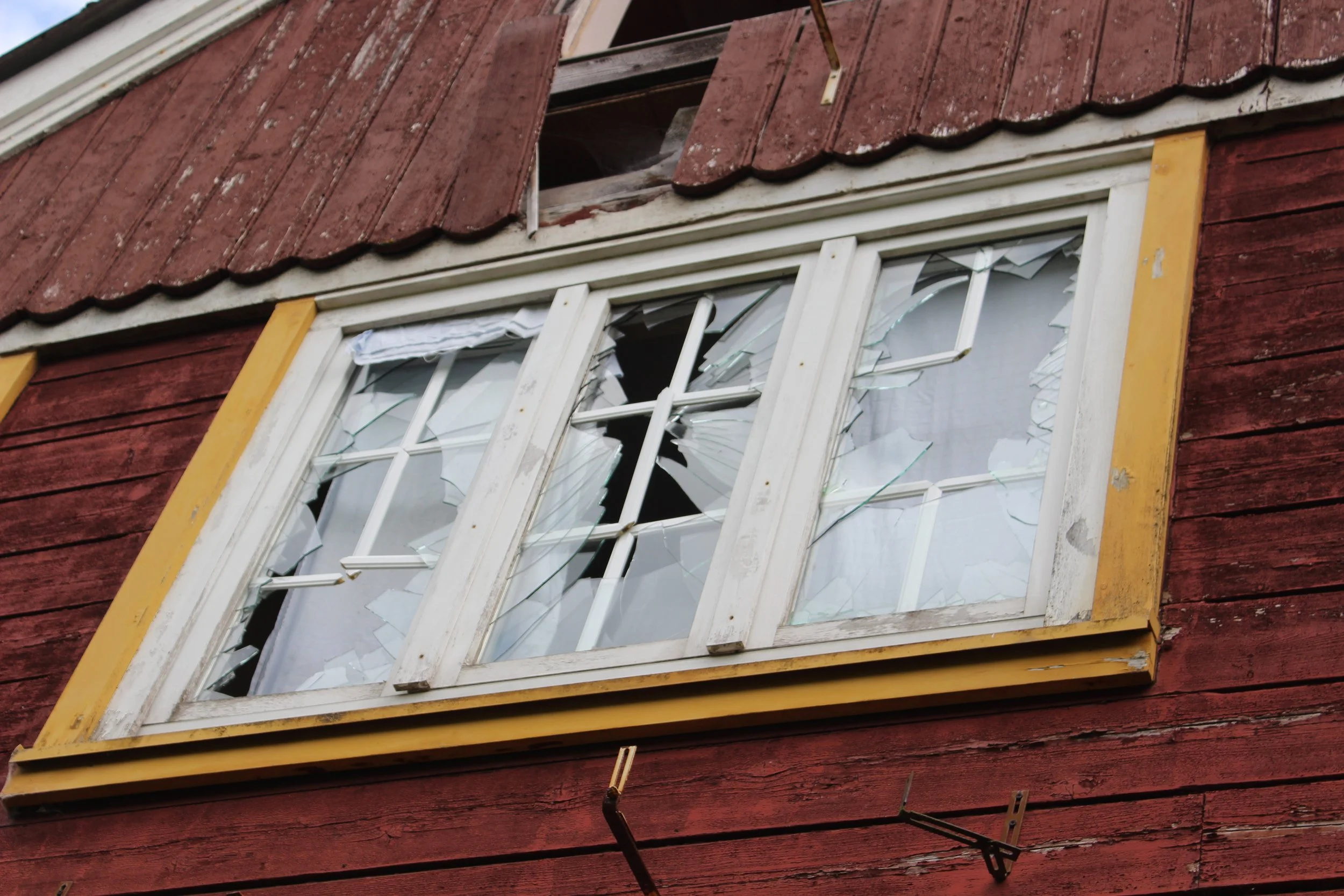 Fenster eines alten Hauses mit zerbrochenem Glas in einem roten Holzhaus, Rahmen in Weiß mit gelber Umrandung.