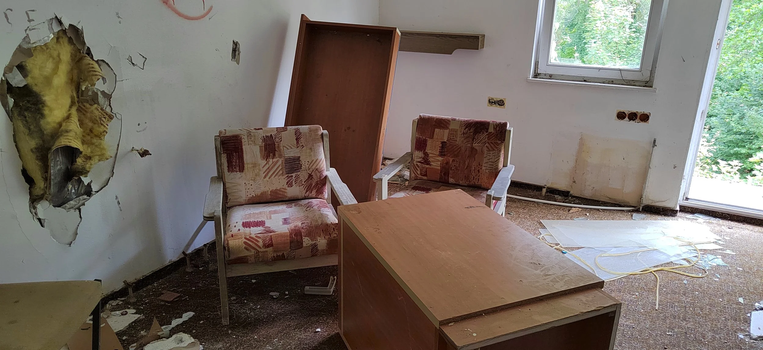 Raum mit beschädter Wand, zerstörtem Boden, zwei alte Sessel, einem umgestürzten Tisch, offener Fenster und sichtbarer Kabeln und Schutt auf dem Boden.