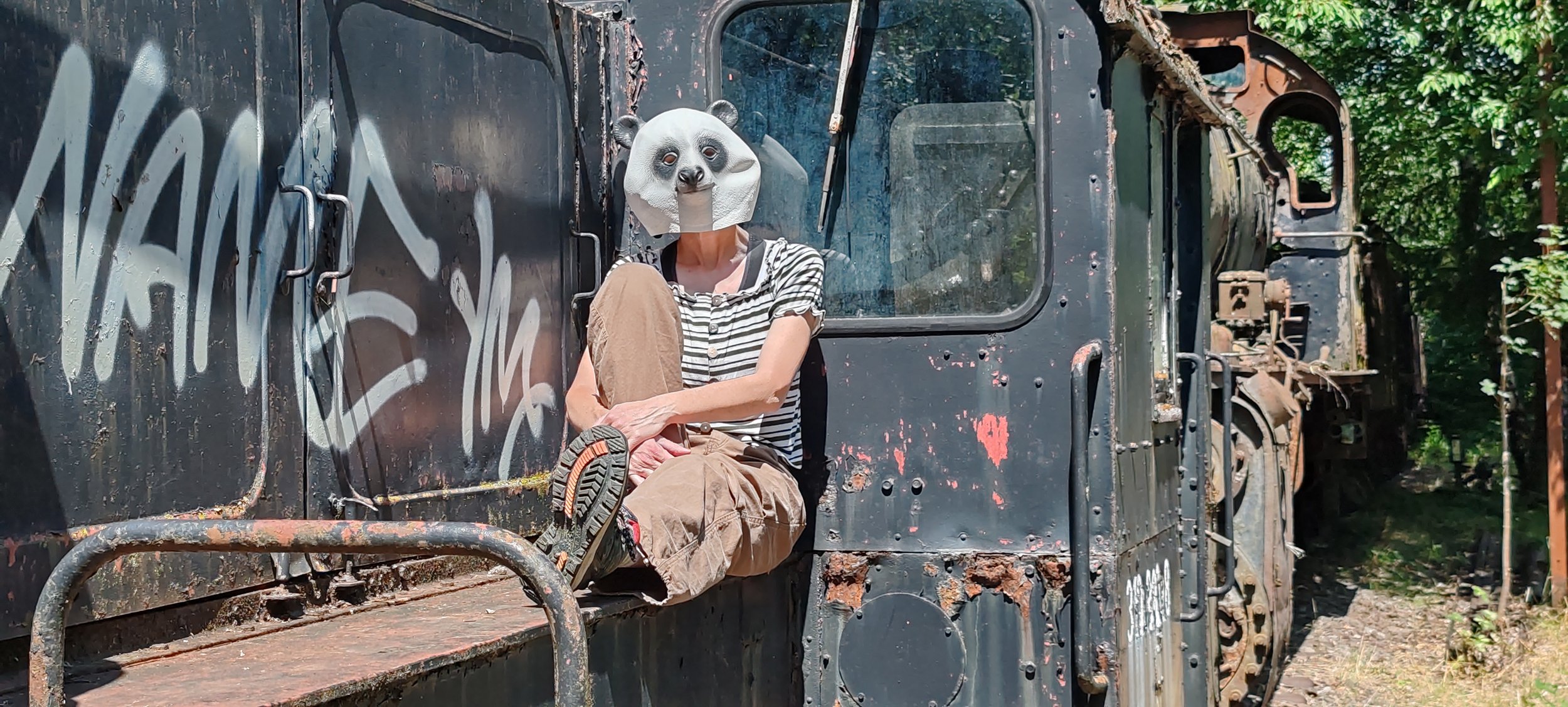 Person sitzt auf einer alten, verrosteten Lokomotive, trägt eine Panda-Maske und einen Streifenkleidung. Umgeben von grünen Bäumen im Freien.
