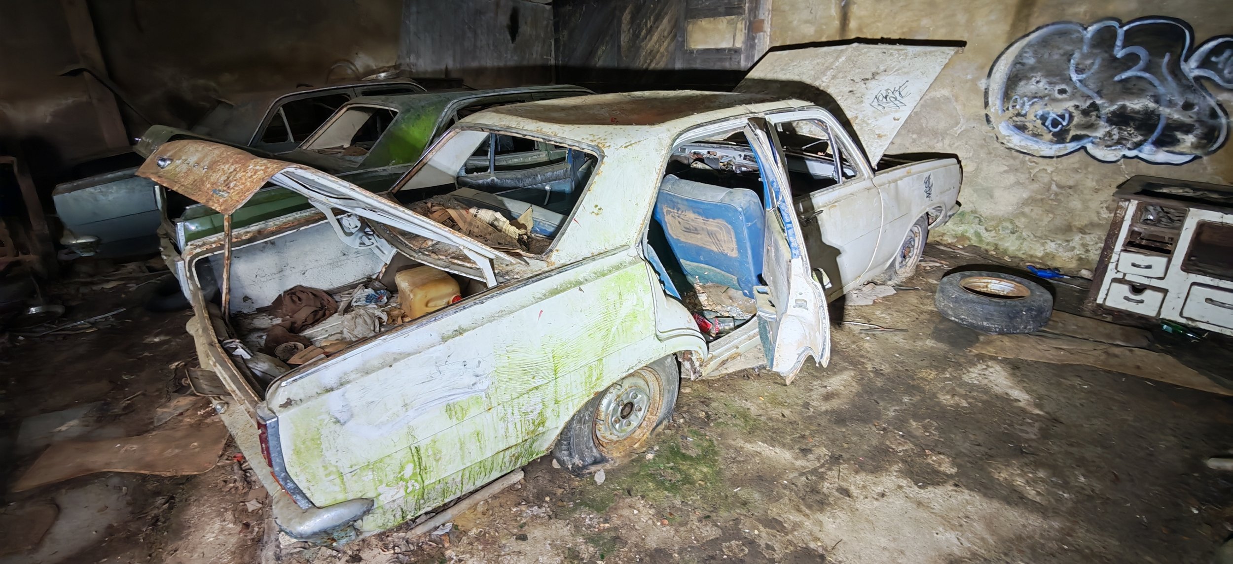 Verrosteter und beschädigter alter Auto in einer dunklen Garage oder Lagerraum, daneben ein Reifen und alte Möbel