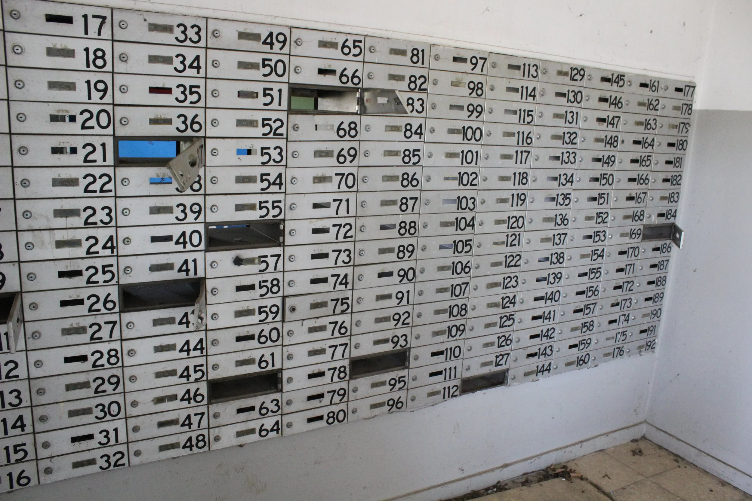 Mehrfamilienbriefkästen mit nummerierten Fächern, einige Fächer sind offen oder beschädigt, an einer weißen Wand montiert.