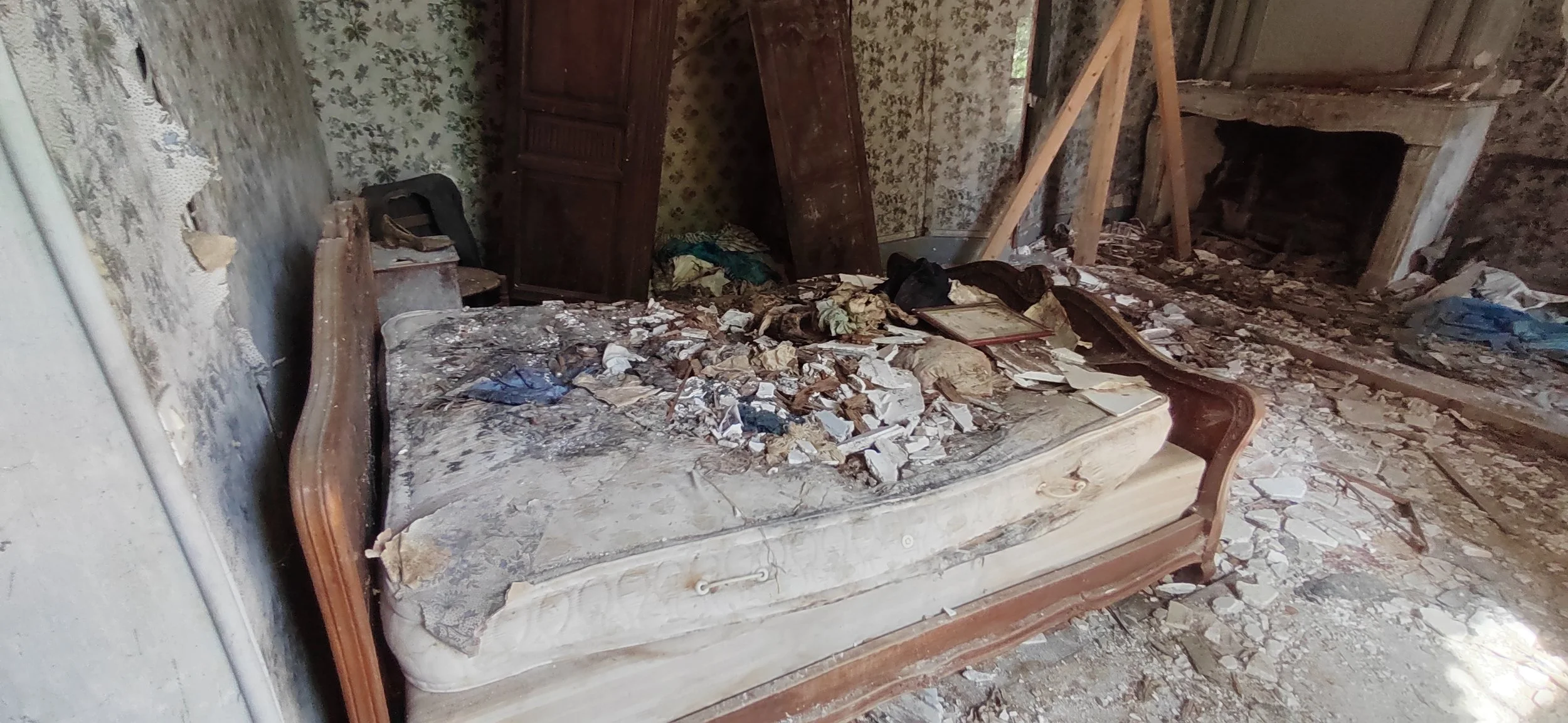 Ein alter, beschädigter und verlassener Raum mit einem zerstörten Bett, das mit Schmutz, Staub und Objekten bedeckt ist, sowie zerbrochenen Wänden und unordentlichem, verfallenem Mobiliar.