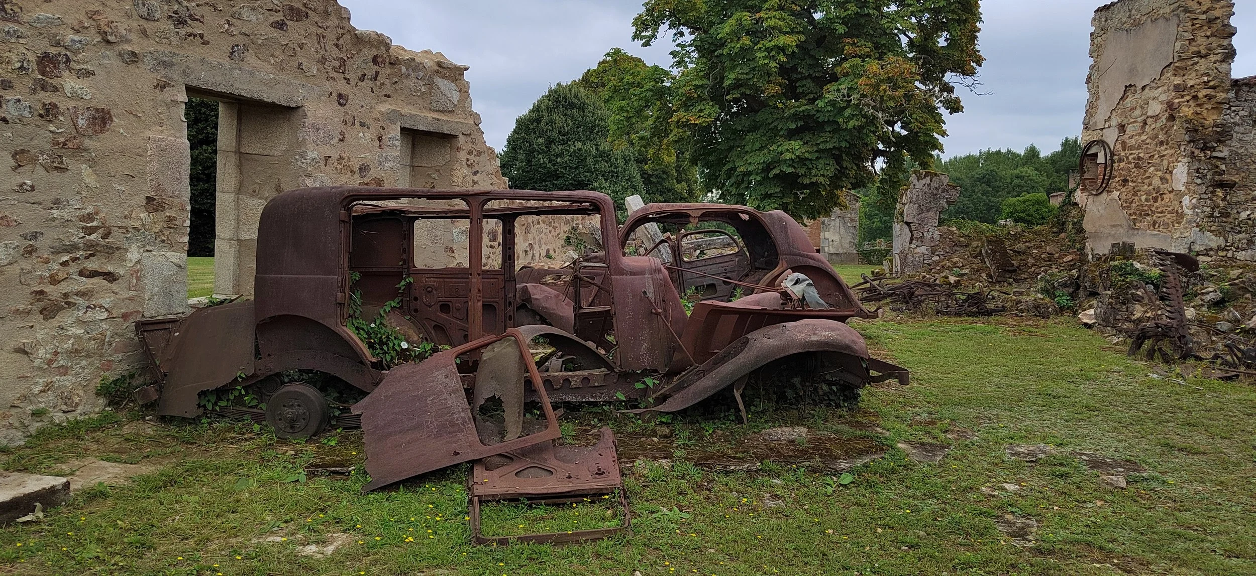 Verrosteter, verlassener Oldtimer-Auto-Rahmen in einer grünen Wiese neben alten, zerfallen Gebäuderuinen, umgeben von Bäumen.