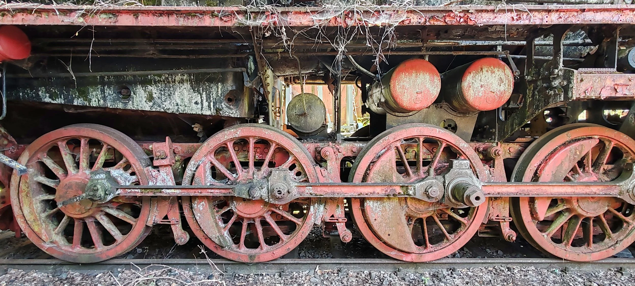 Alte, verrostete Dampflokräder und Antriebsteile auf Schienen, teilweise mit Moos und Staub bedeckt.
