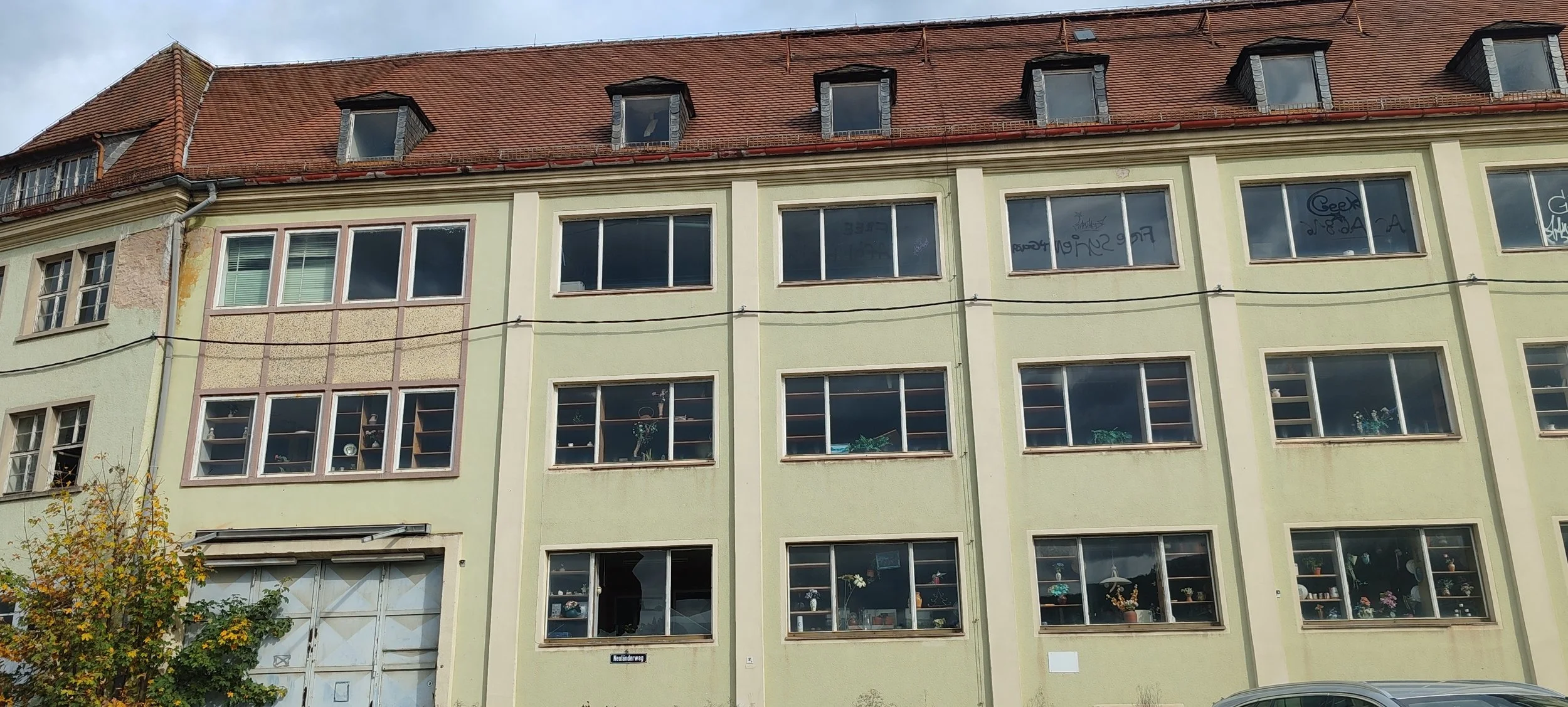 Mietshaus mit mehreren Fenstern an einer grünen Fassade, Dach mit roten Ziegeln, Bäume im Vordergrund.