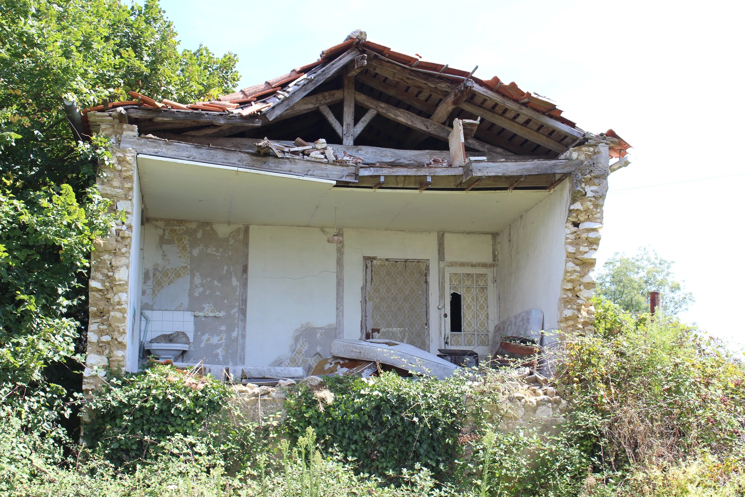 Ein verfallenes, halboffenes Haus mit beschädigtem Dach, umgeben von Büsche und Bäumen.