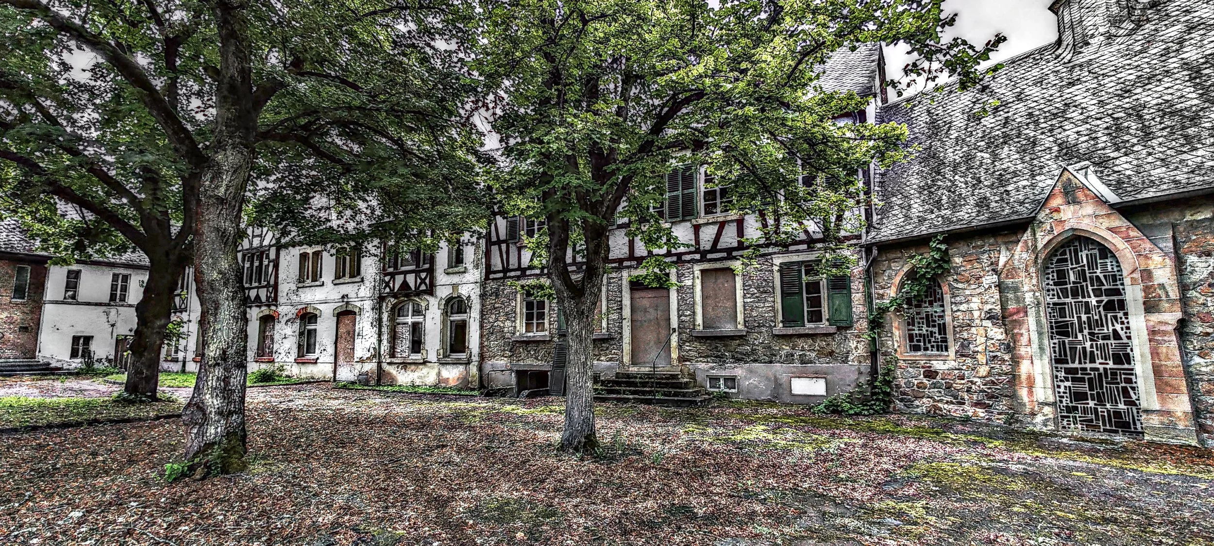 Historisches Gebäude mit einer großen Baum im Vorgarten, im Stil eines alten, steinernen Hauses mit Fensterläden und Zierfenstern, umgeben von Bäumen und Laub auf dem Boden.