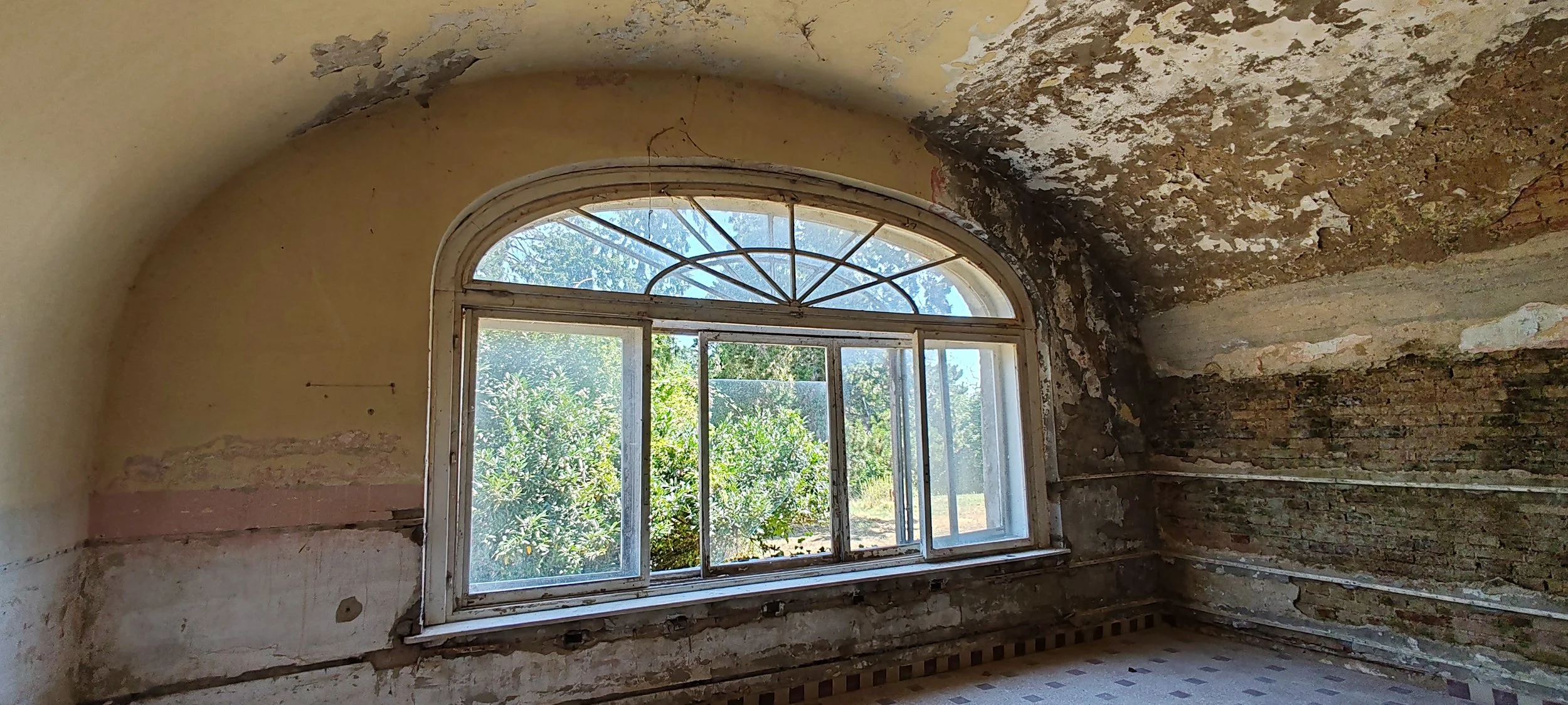 Ein altes, verfallenes Zimmer mit einer großen, halb geöffneten Fensterfront, durch die man Garten und Bäume sieht, die Decke ist schmutzig und beschädigt, die Wände sind brüchig und teilweise offen, der Boden ist mosaikartig gefliest.
