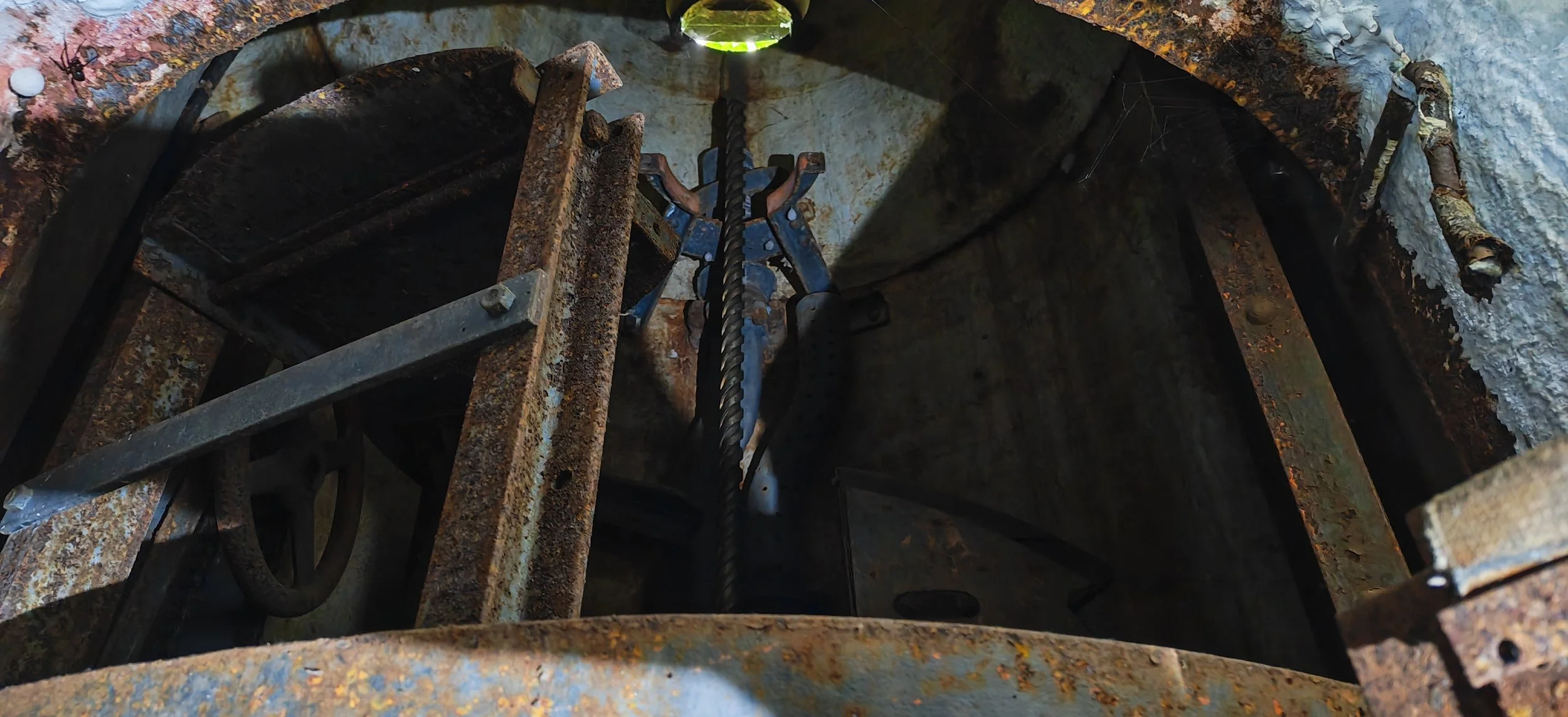 Blick in eine alte, rostige Turmuhr mit mechanischen Teilen und einem Zeiger im oberen Bereich.