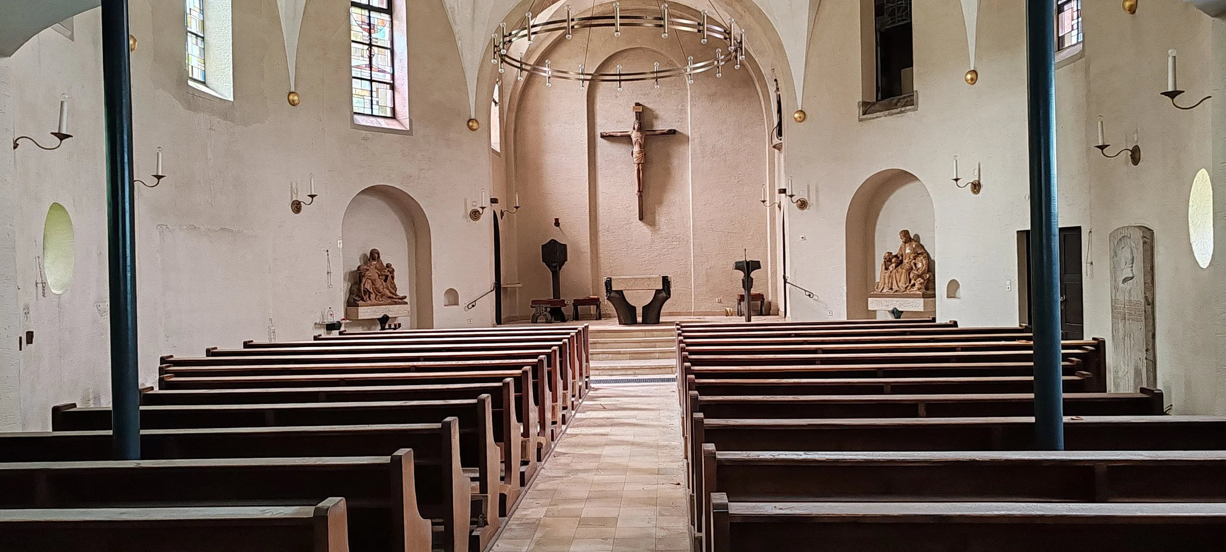 Ein leeres Kircheninnere mit Holzbänken, einem Altar, einer Kreuzfigur, Kerzenleuchtern an den Wänden und bunten Fenstern.